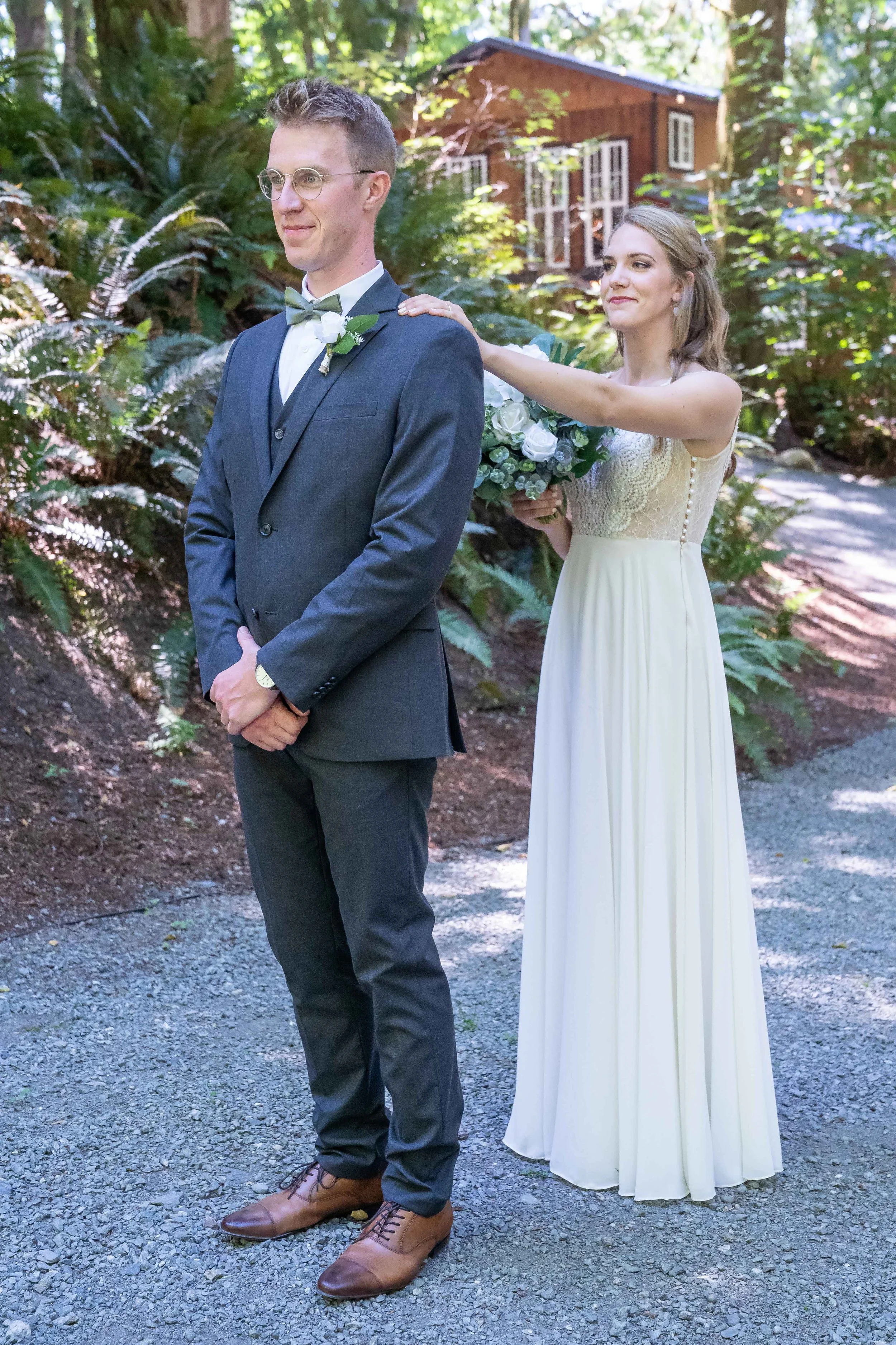 Photo of a first look at a wedding in a fern-covered venue in Skagit County, Washington. The bride is standing behind the groom with her hand on his shoulder.