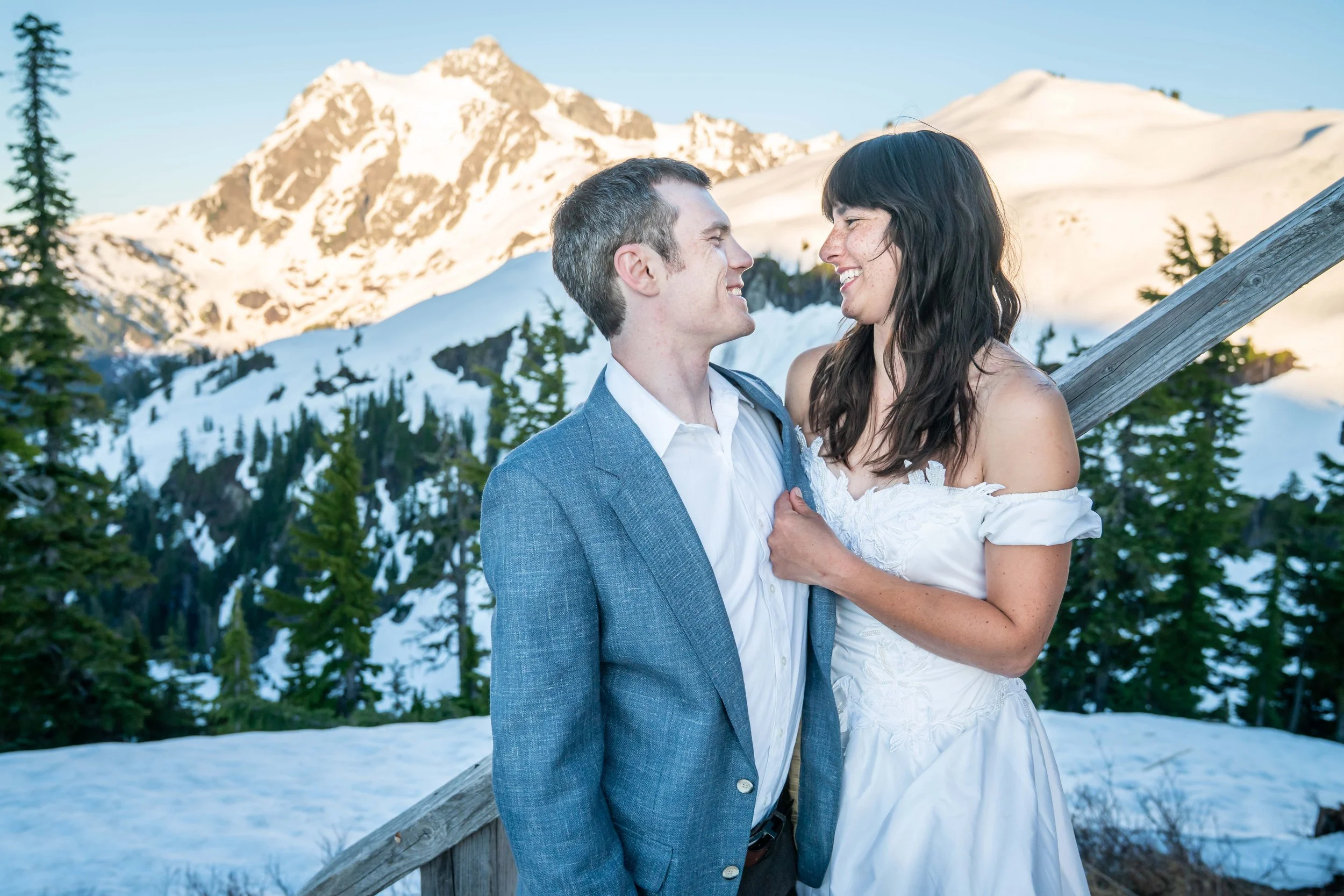 A photo of a couple in a suit and wedding dress standing together and smiling at each other. There's snow around them and Mt. Shuksan in the background.
