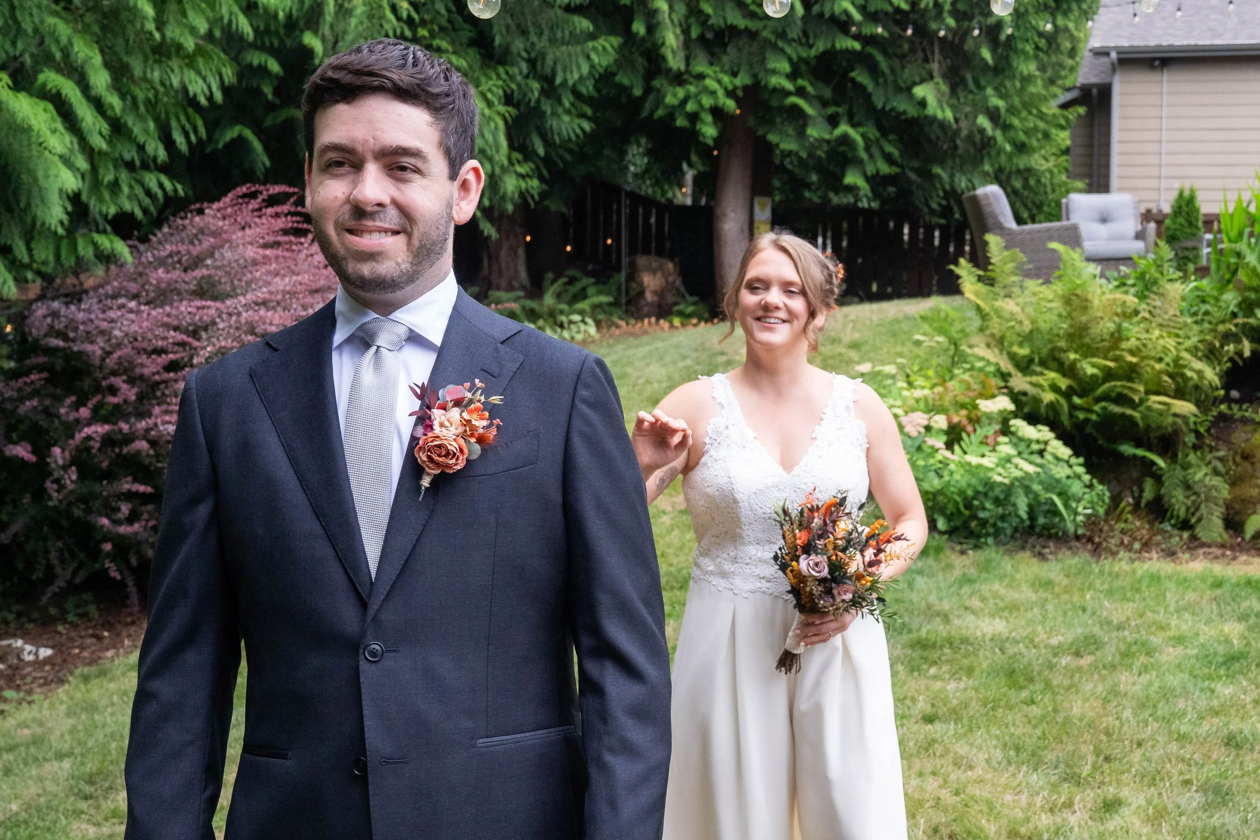 Photo of a first look in a forested backyard with the bride walking up behind the groom and reaching out to touch his shoulder. The bride is holding a bouquet and the groom is in a dark suit.