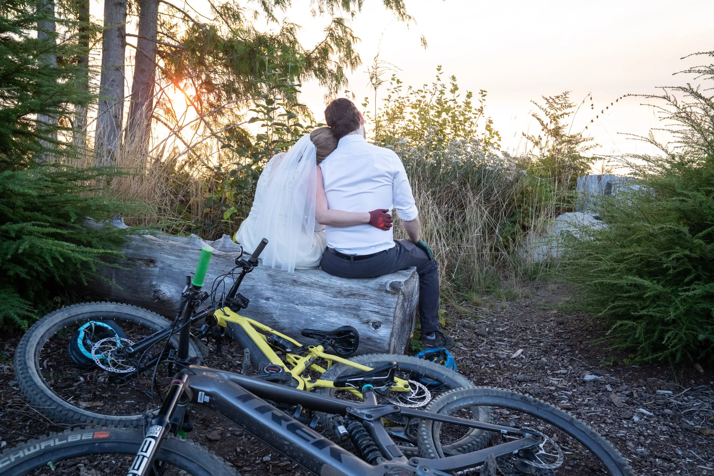 A photo of a bride and groom in wedding clothes and mountain bike gear sitting on a log and watching the sunset from Galbraith Mountain. Their mountain bikes are laying on the ground in the foreground.