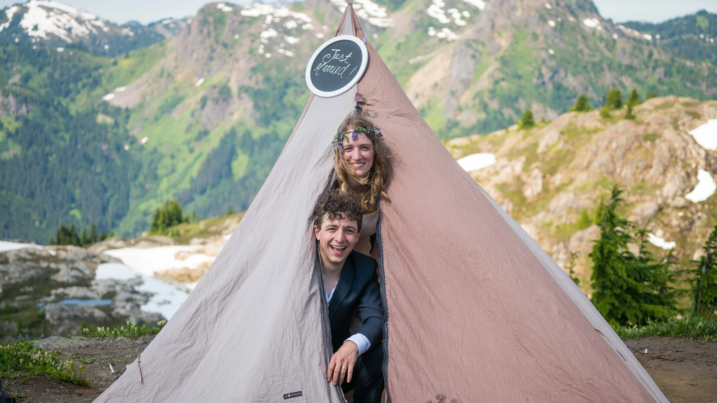 A bride and groom sticking their heads out of a tent after waking up in the North Cascades during a backpacking elopement. There's a "Just Married" sign on the tent and the mountains at sunrise are visible behind them.