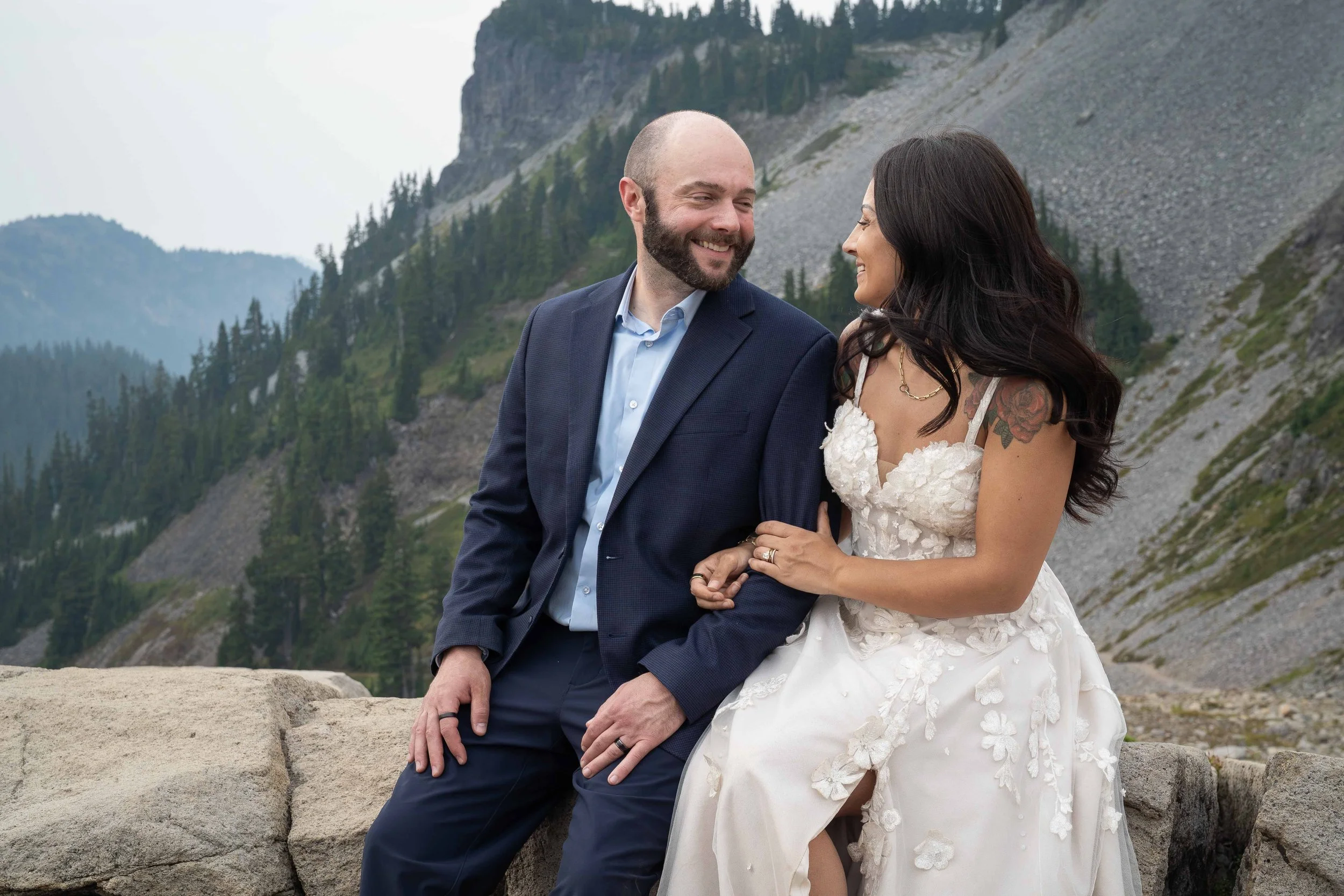 A bride and groom in wedding clothes sit on a boulder in front of Table Mountain and smile at each other during their Ptarmigan Ridge elopement.