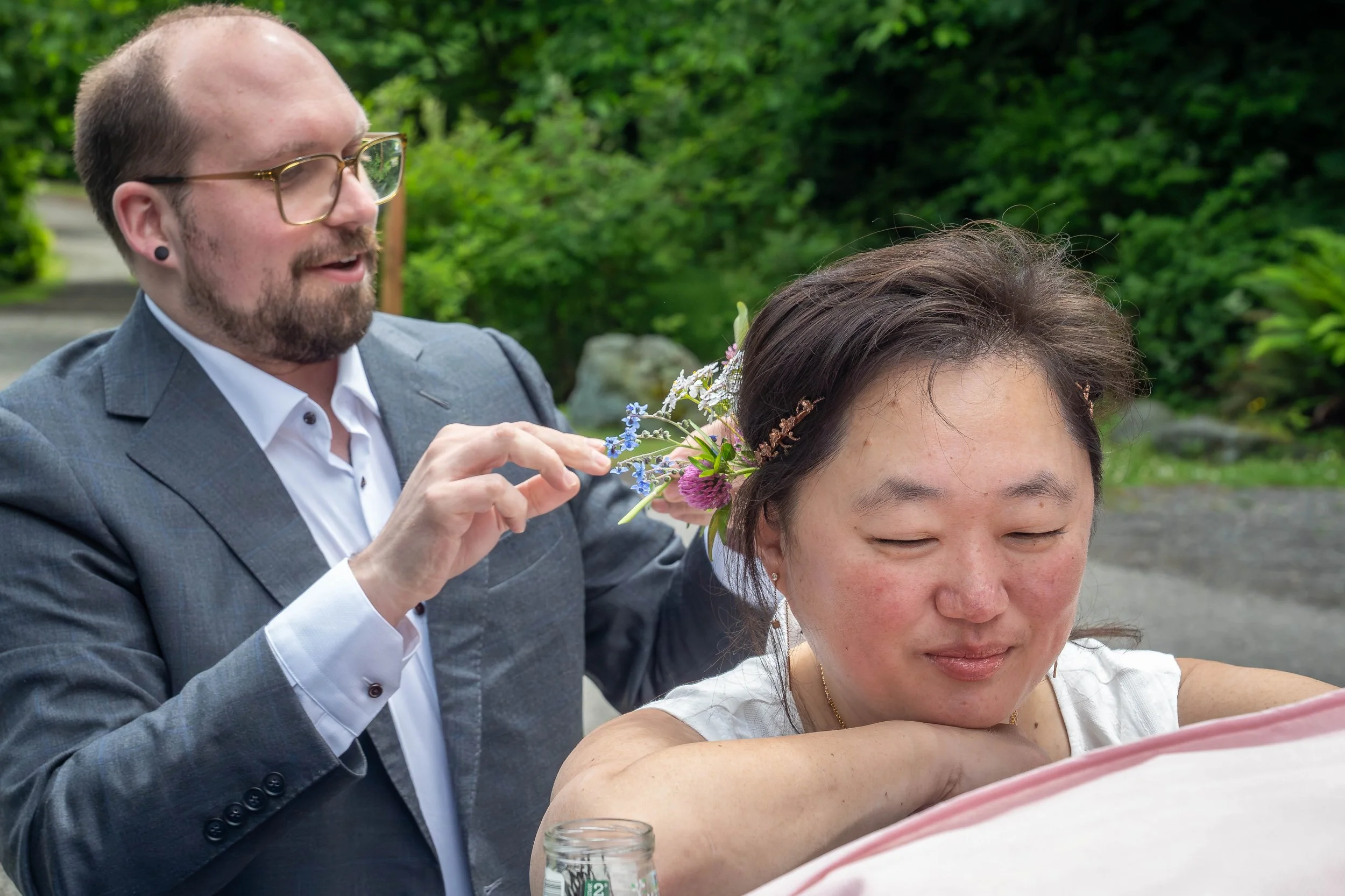 A bride is smiling and looking down while a hairdresser in a suit pins flowers into her hair. Taken during an elopement photography session in Larrabee State Park.