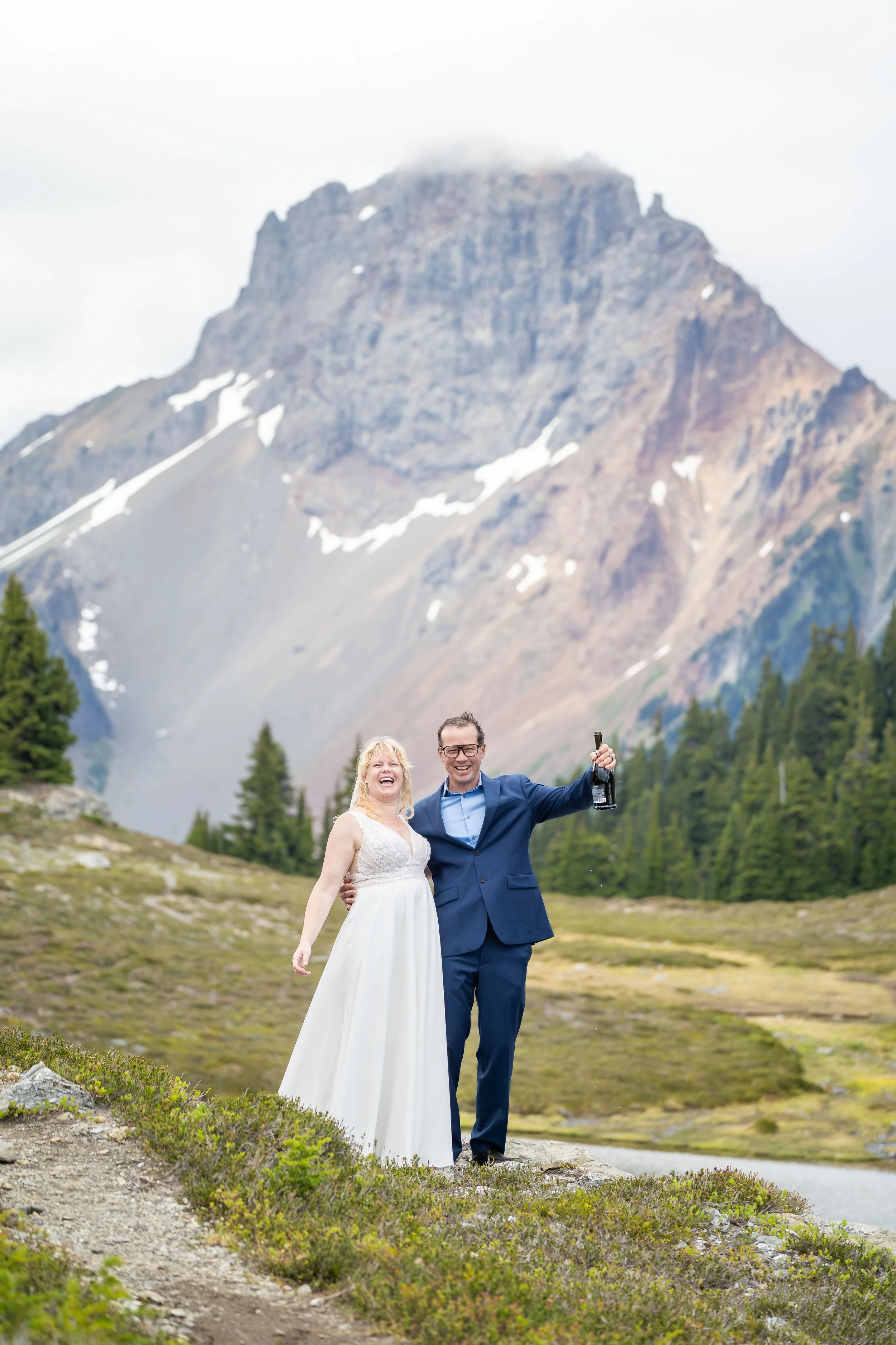 A bride and groom celebrate with a bottle of champagne after their Yellow Aster Butte hiking elopement. They are standing in a subalpine meadow next to an alpine lake, with American Border Peak visible behind them.