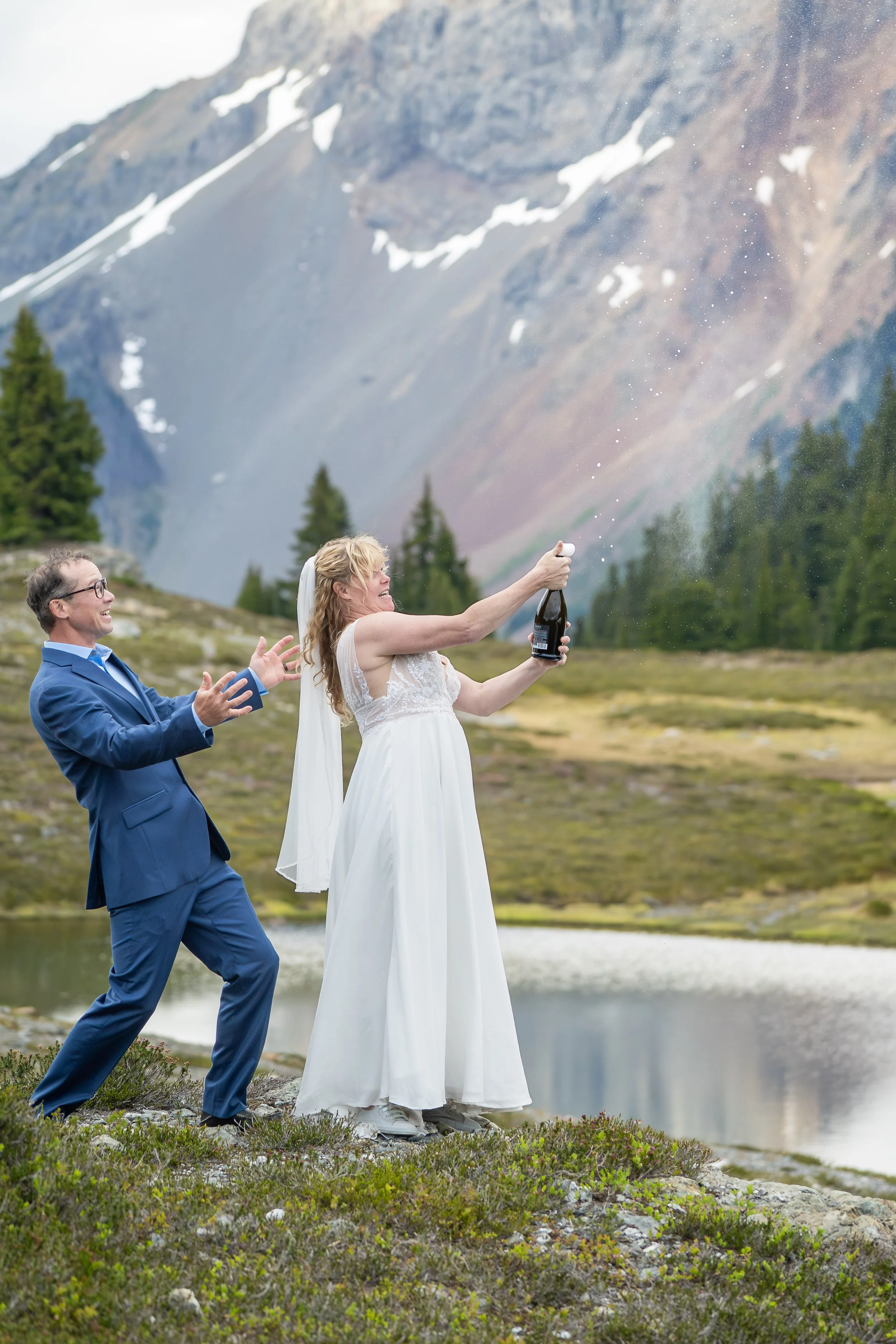 A bride in a wedding dress and a groom in a suit popping a bottle of champagne during their North Cascades elopement, with American Border Peak visible behind them. They are standing in a subalpine meadow in front of a snow-fed alpine lake.