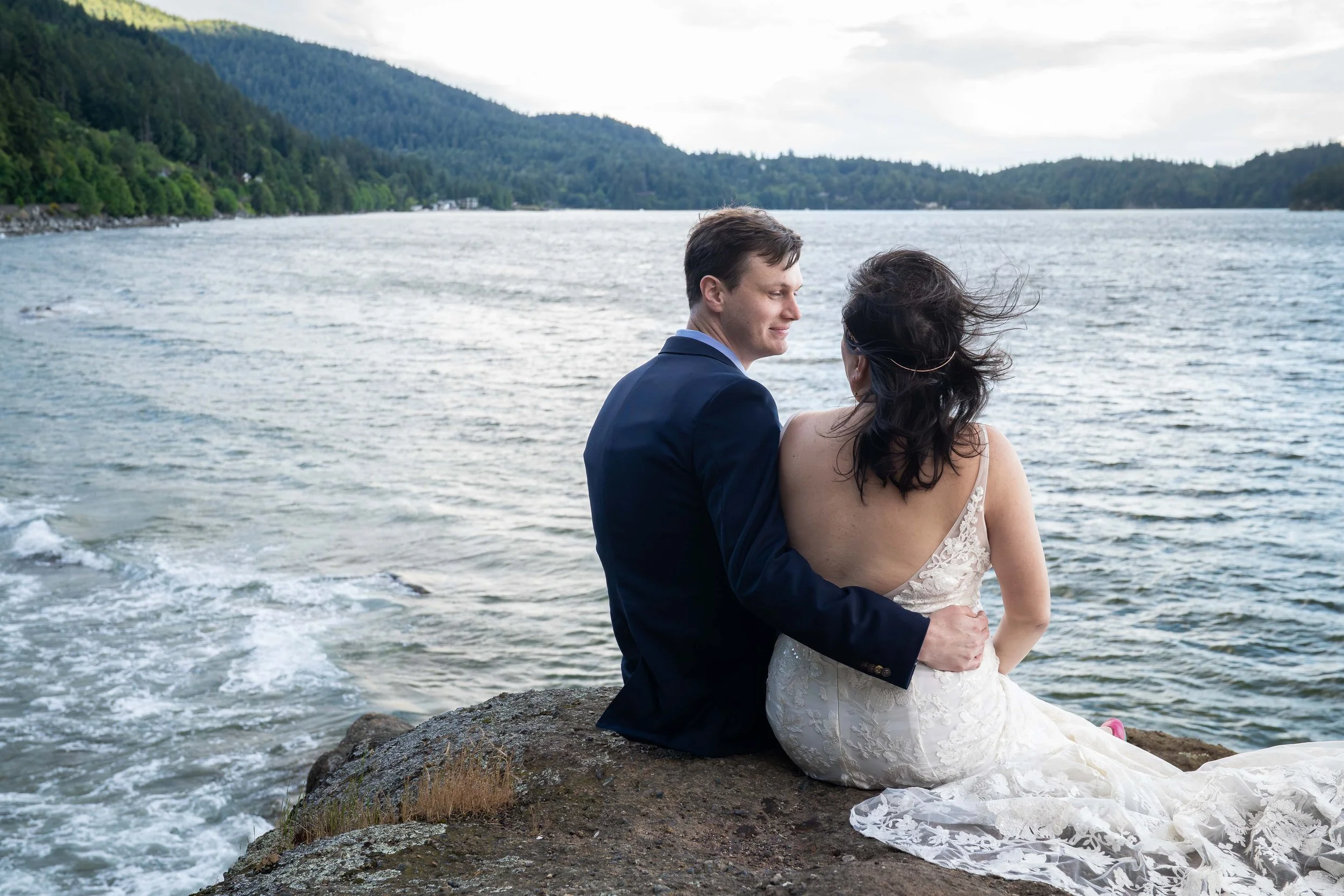 A bride and groom sit together on a rocky bluff overlooking the water during sunset at Teddy Bear Cove in Bellingham, Washington.