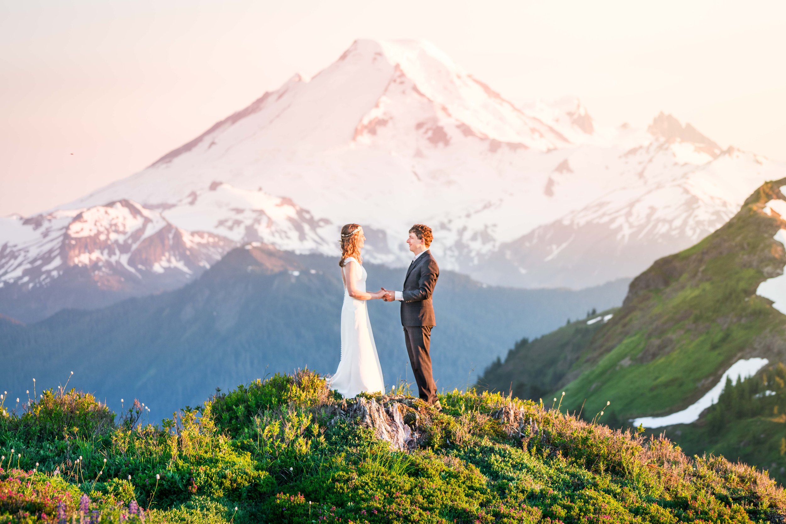 A bride and groom in wedding clothes standing in front of each other and holding hands. They're standing on Yellow Aster Butte, with a snowy Mount Baker directly behind them.