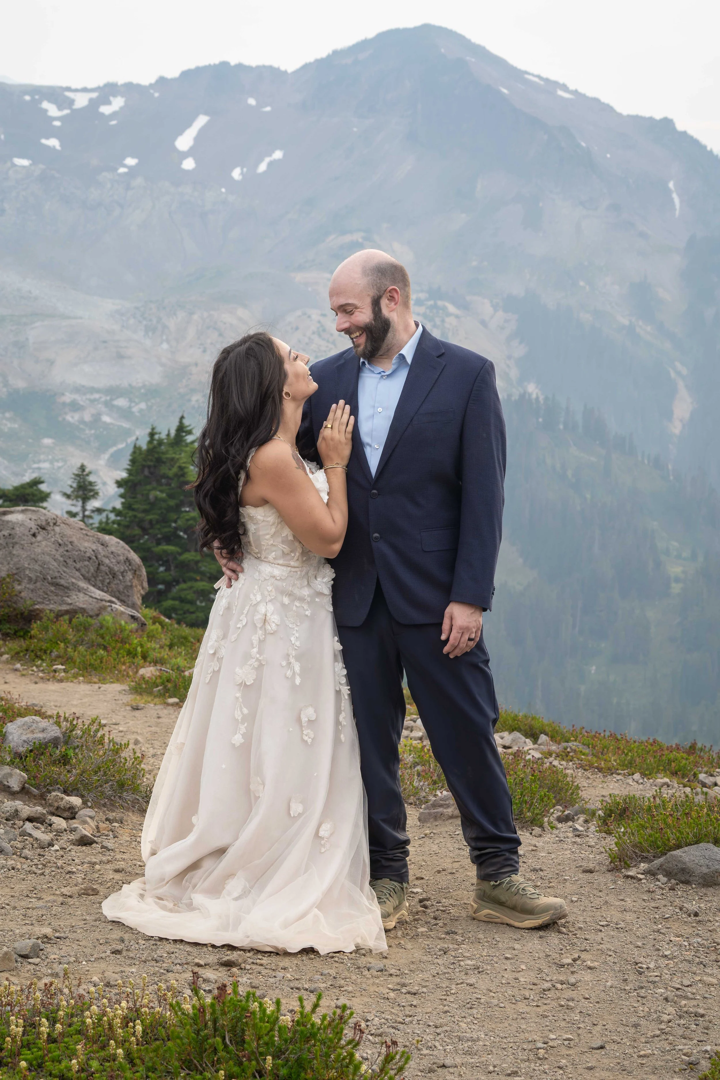A bride and groom laughing with one another during their Ptarmigan Ridge elopement near Mt. Baker. They are standing on a trail surrounded by blooming heather with the slopes of Mt. Baker in the background.
