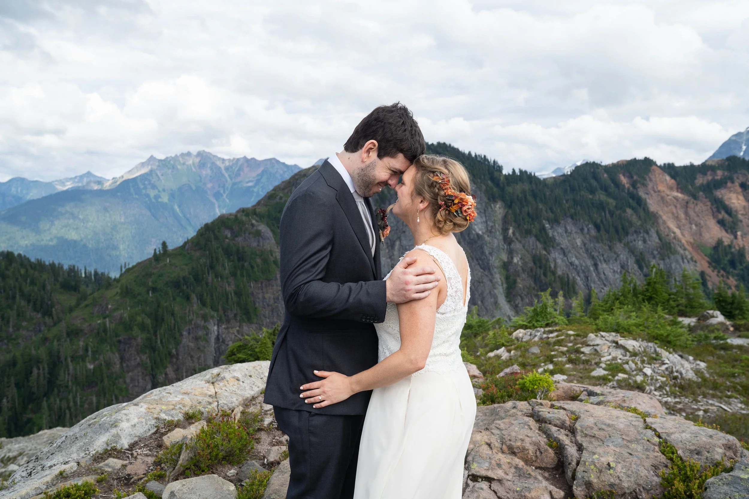 Photo of a bride and groom eloping at Artist Point, with the North Cascades behind them. The couple is in wedding attire and they are holding each other with their foreheads pressed together.