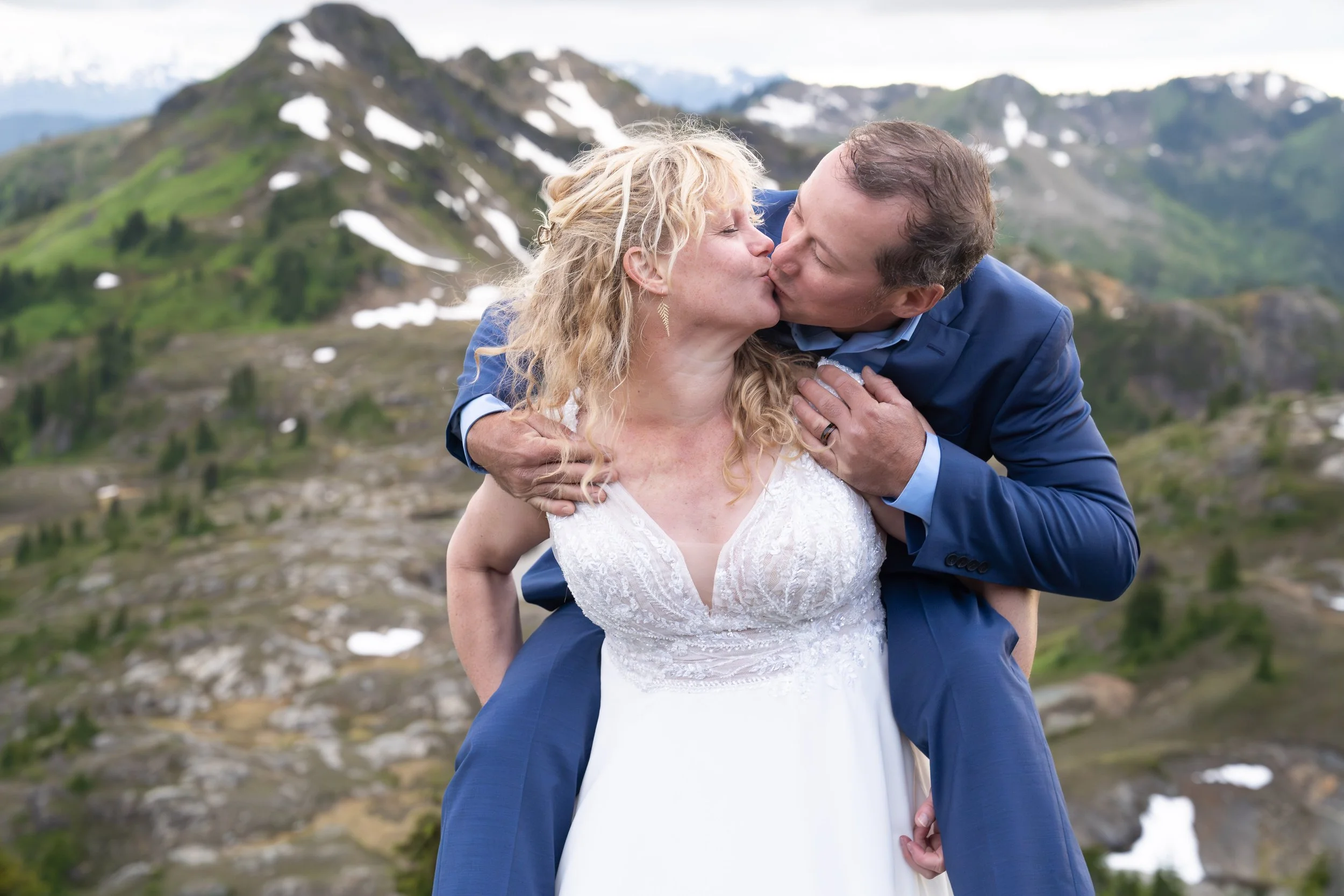 A bride holds her groom on her back during a North Cascades elopement at Yellow Aster Butte. They are kissing and the groom's ring is clearly visible. Sunset is taking place over Keep Kool Butte and Mount Baker behind them.