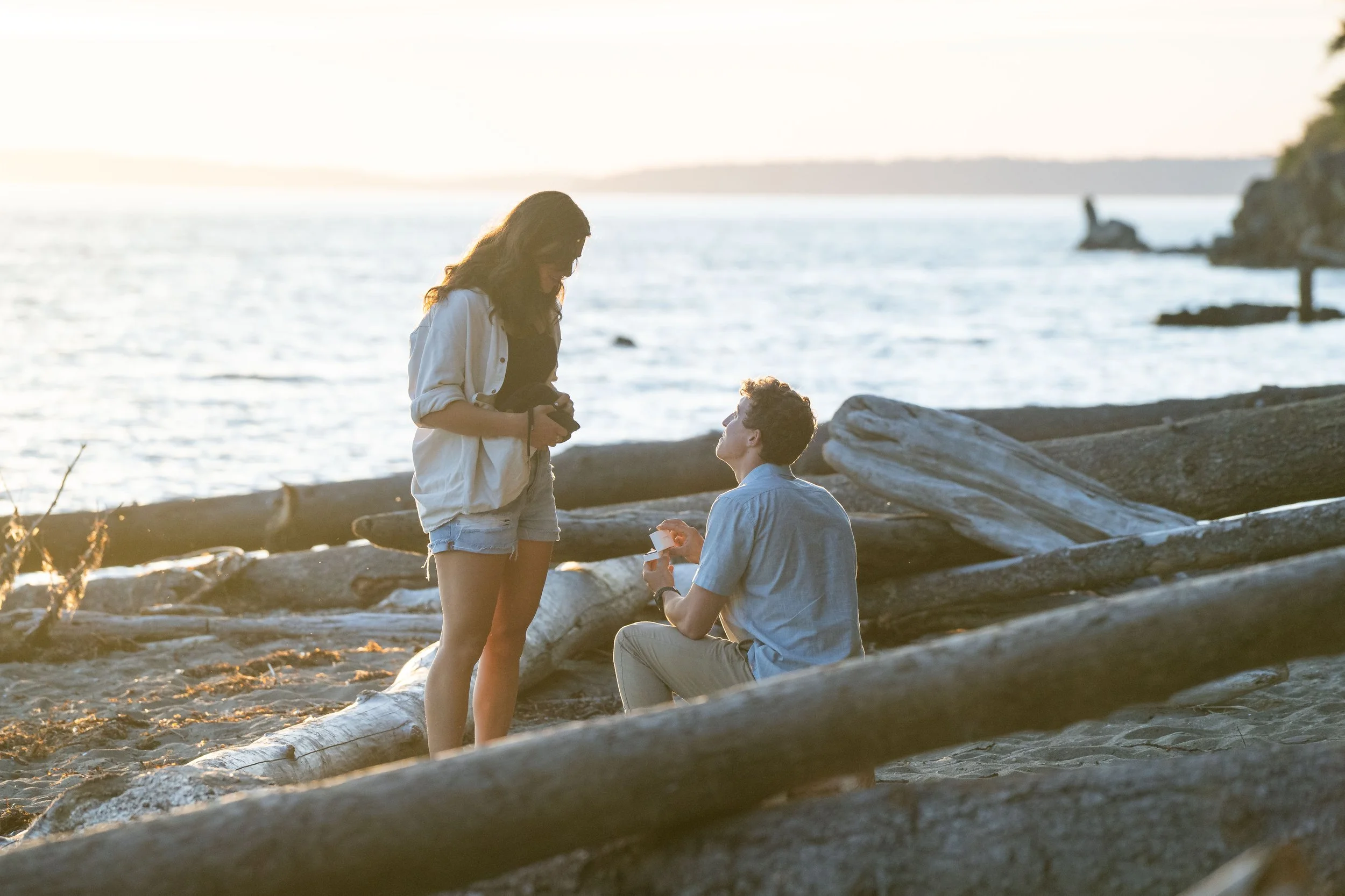 A photo of a young man down on one knee and holding a ring box as he proposes to his girlfriend. They're on the beach at Clayton Beach, framed by driftwood and the water at sunset.