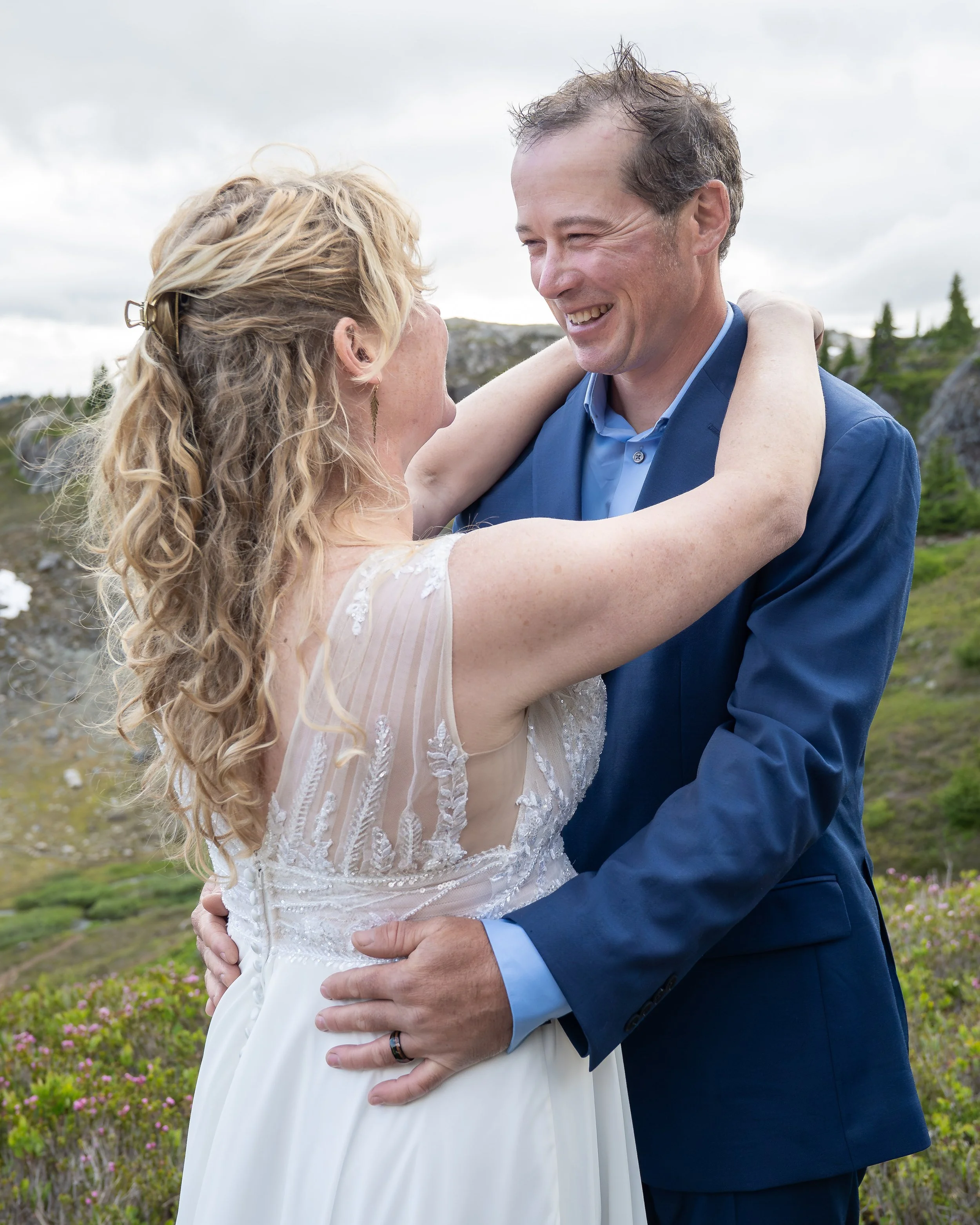 A bride and groom holding each other and smiling during their elopement ceremony at Yellow Aster Butte, in the North Cascades, Washington. They are standing in a subalpine meadow surrounded by blooming heather and wildflowers.