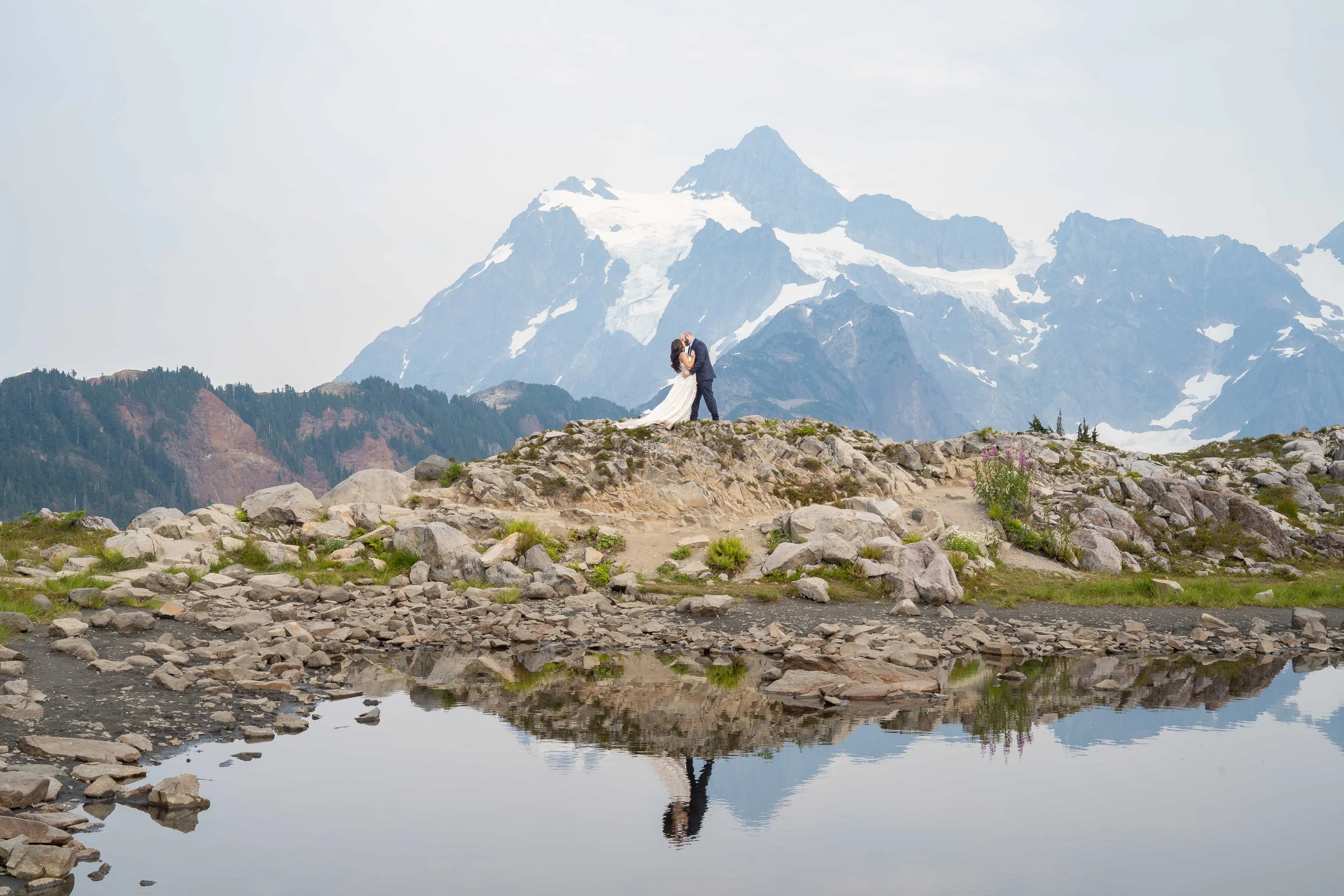A bride and groom kissing in front of Mt. Shuksan during a Mt. Baker elopement at Artist Point. They're standing on a rocky pedastal above an alpine lake with their reflection mirrored in the lake.
