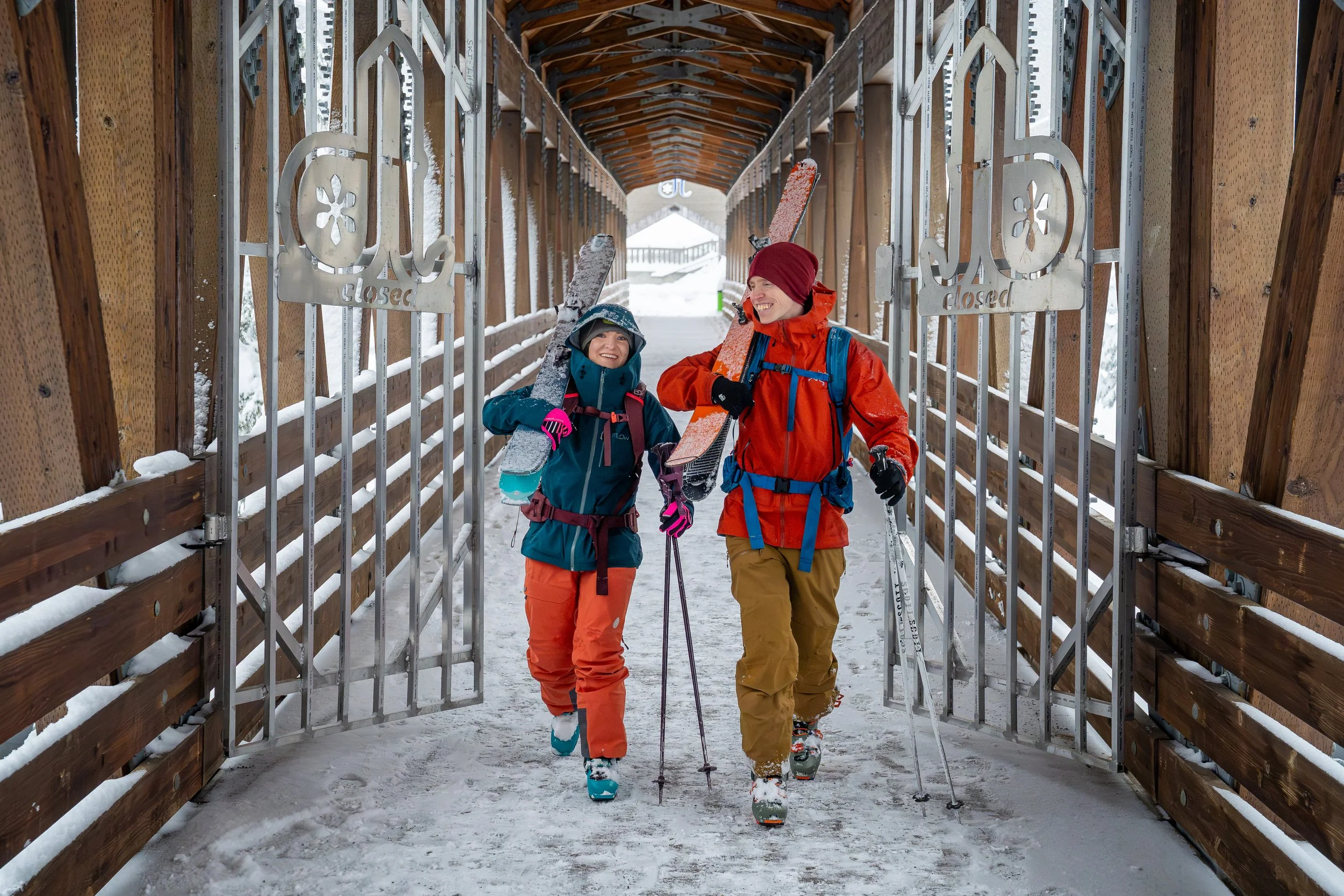 A photo of a couple in ski clothes with skis over their shoulder smiling as they walk towards the camera together. They're on a wooden bridge at Alpental and walking through large metal gates with the Alpental logo.
