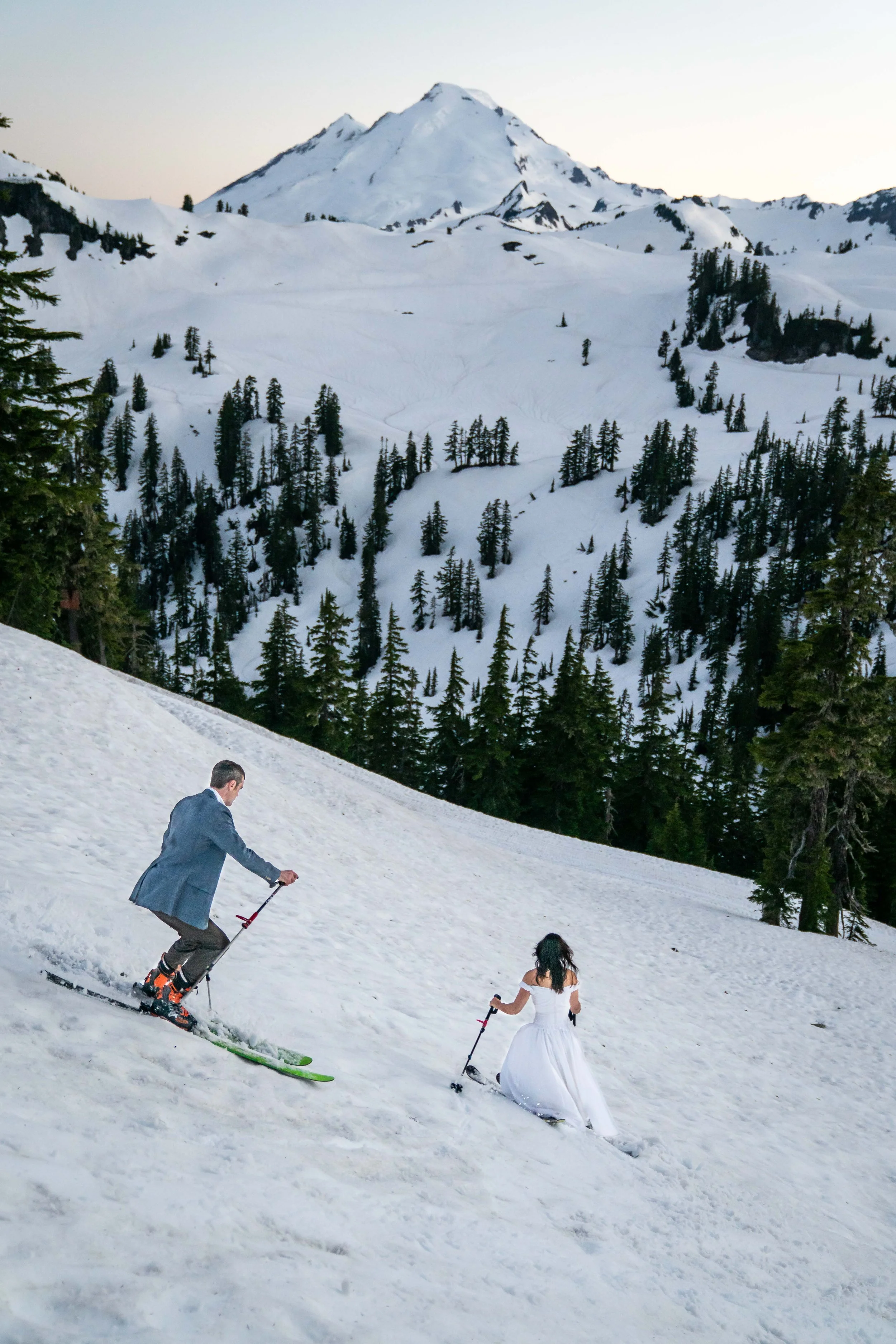 A photo of a woman in a wedding dress and a man in a suit skiing downhill in a snowy landscape, with Mount Baker in the background.