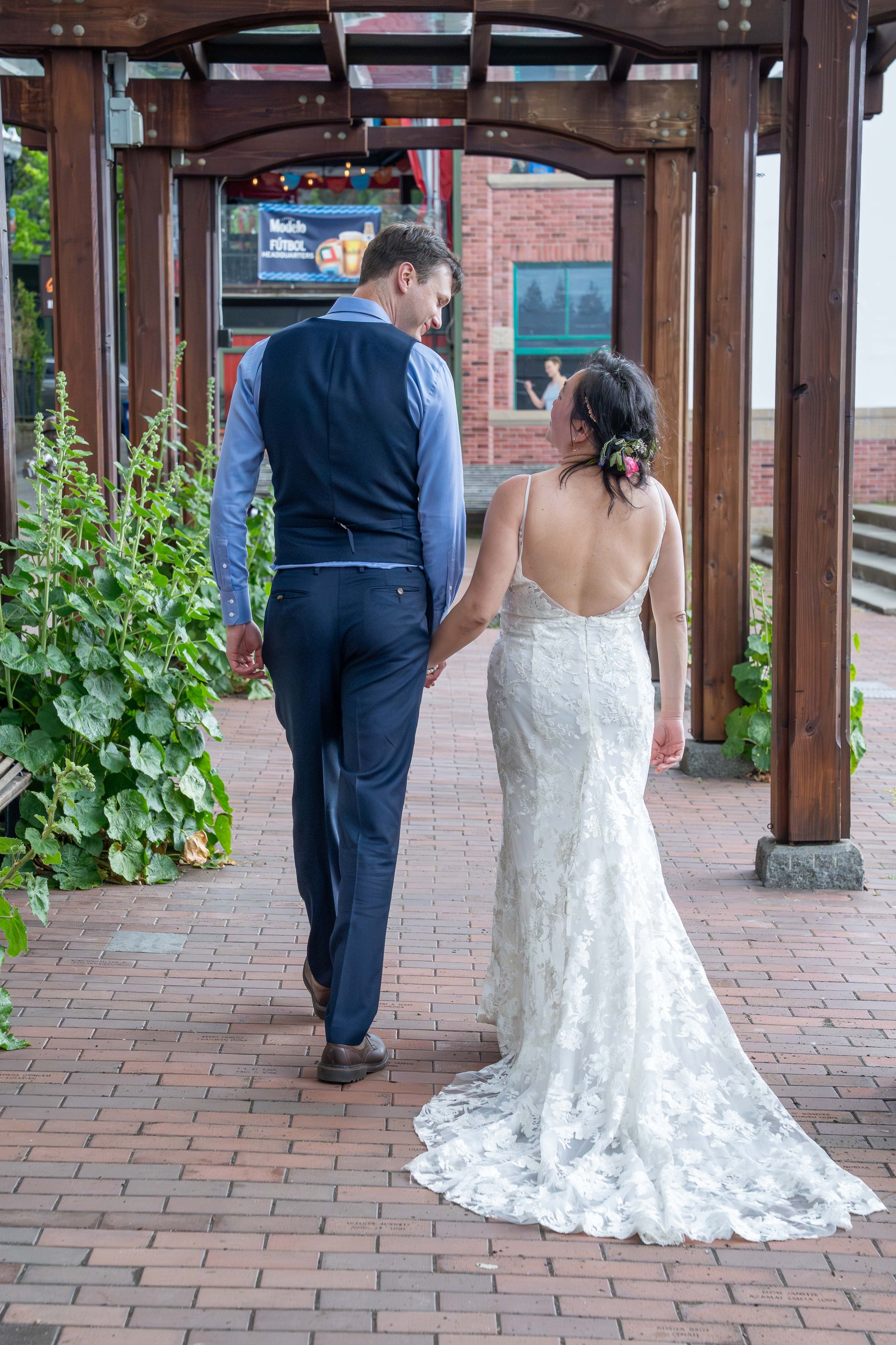 A bride and groom walk away from the camera down a brick-lined plaza in the Village Green in historic downtown Fairhaven after their Bellingham elopement ceremony and first dance.