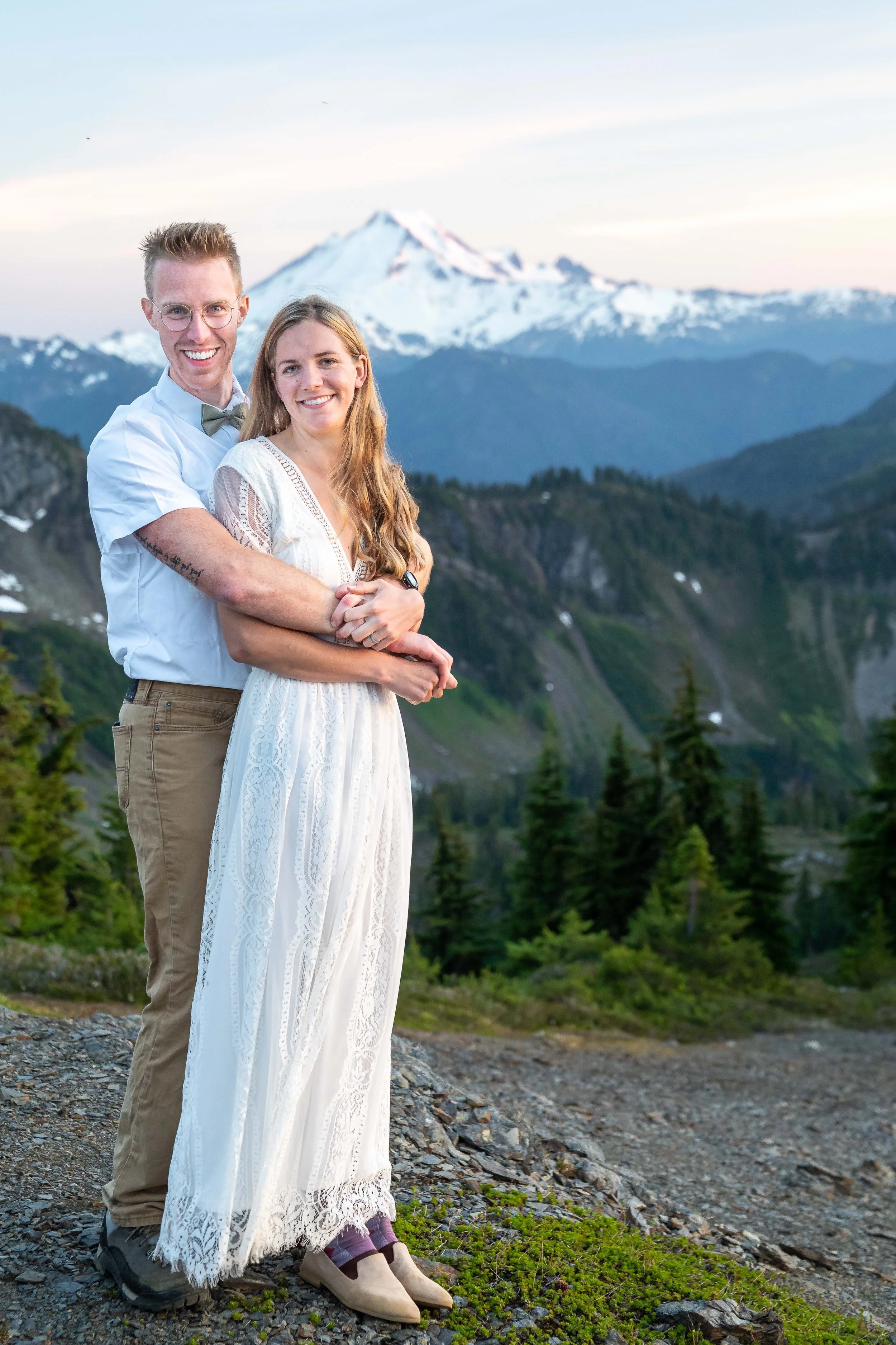 A photo of a bride in a wedding dress and a groom in a bowtie during a backcountry wedding at High Pass in the North Cascades in Washington. Mt. Baker is visible behind them at sunset.