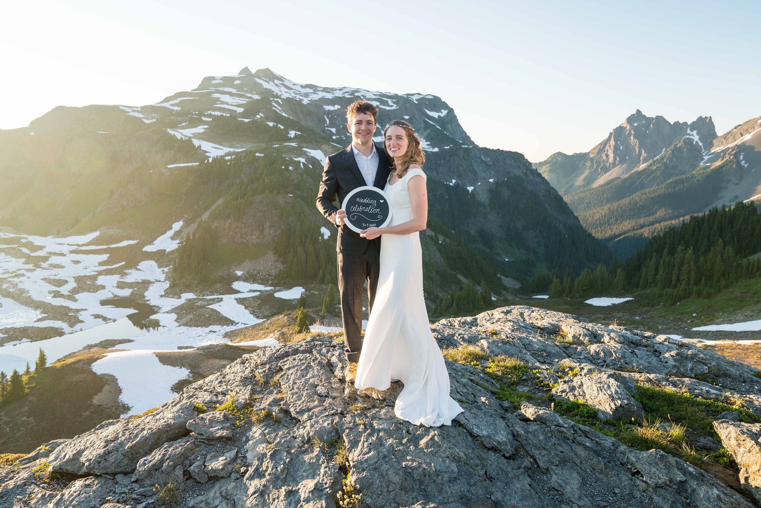 A bride and groom standing together in the North Cascades at sunset with Tomyhoi Peak and American Border Peak behind them. They're holding a sign that says "Wedding Celebration" and there's snow visible on the slopes behind them.