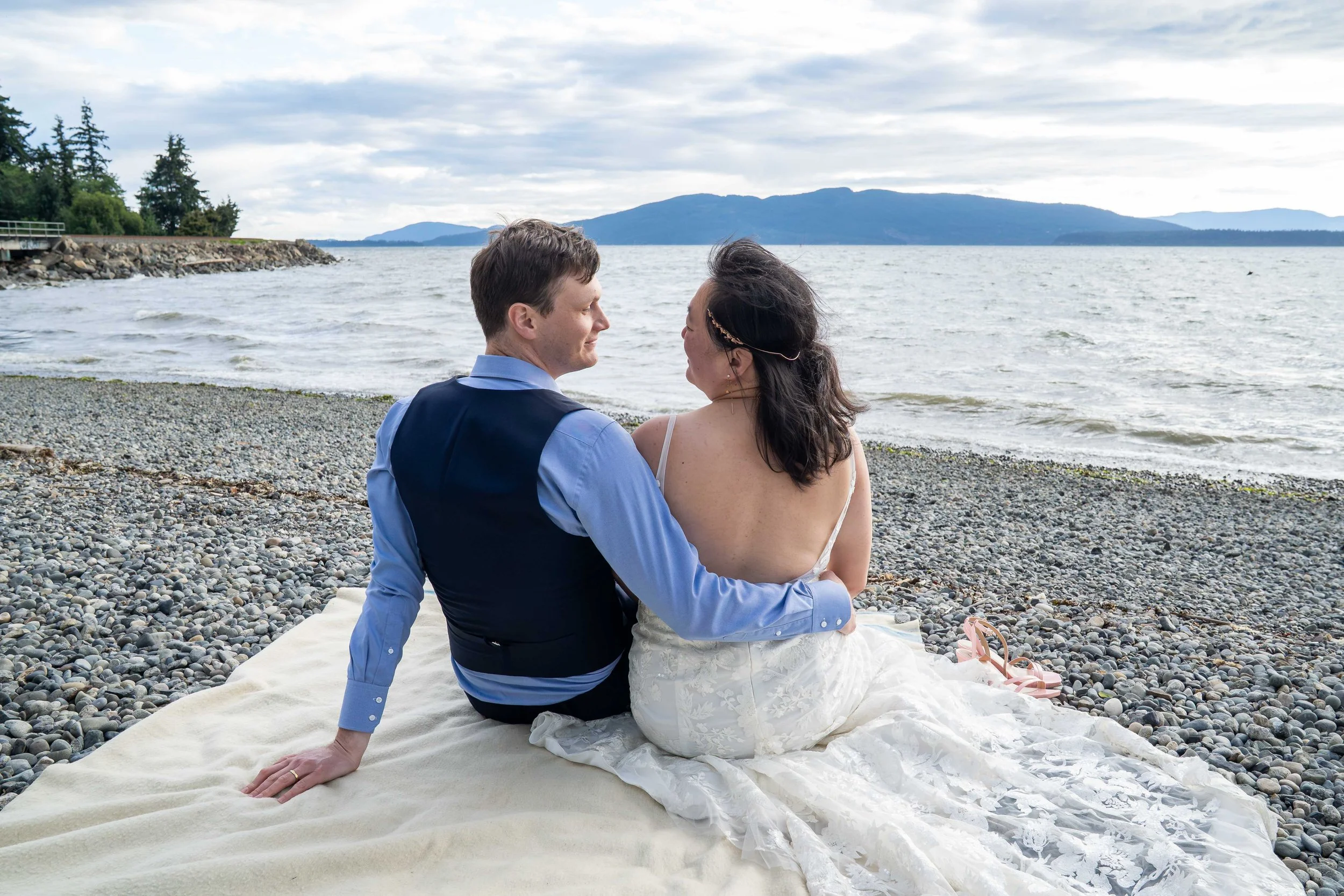 A photo of a bride and groom sitting together on a blanket on the rocky beach at Marine Park after their Bellingham elopement. They are smiling at each other and watching the sunset over the San Juan Islands.