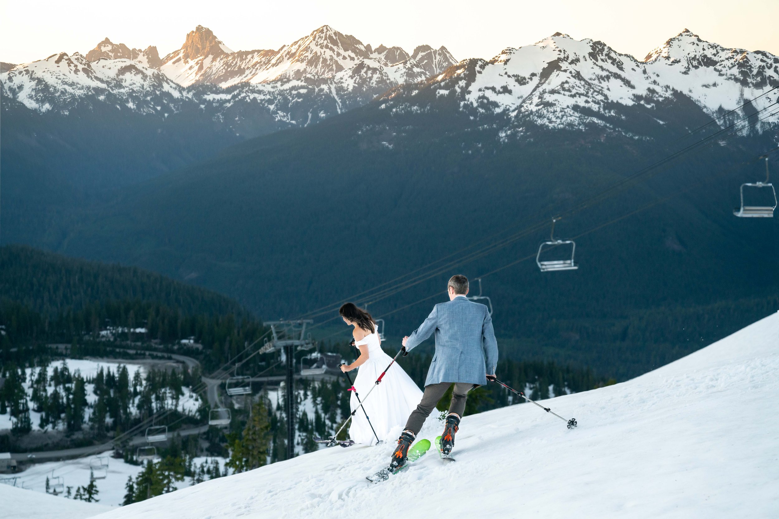 A bride and groom celebrating their anniversary by backcountry skiing in the spring near the Mt. Baker Ski Area at sunset with the North Cascades in front of them.