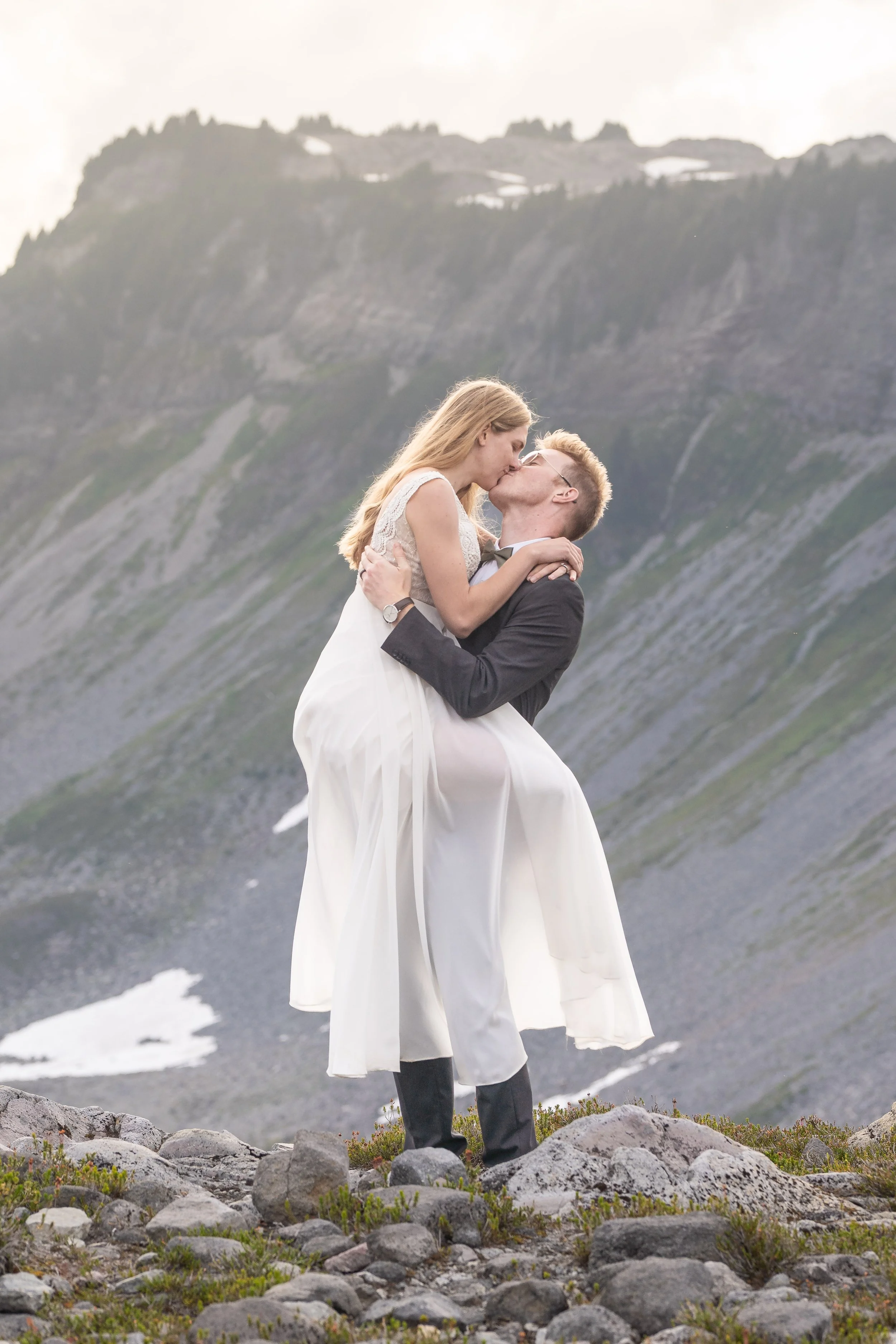 A photo of a bride in a wedding dress and groom in a suit kissing in front of Table Mountain at sunset. The photo is taken from Artist Point. The bride has her legs wrapped around the groom and her arms around his neck.