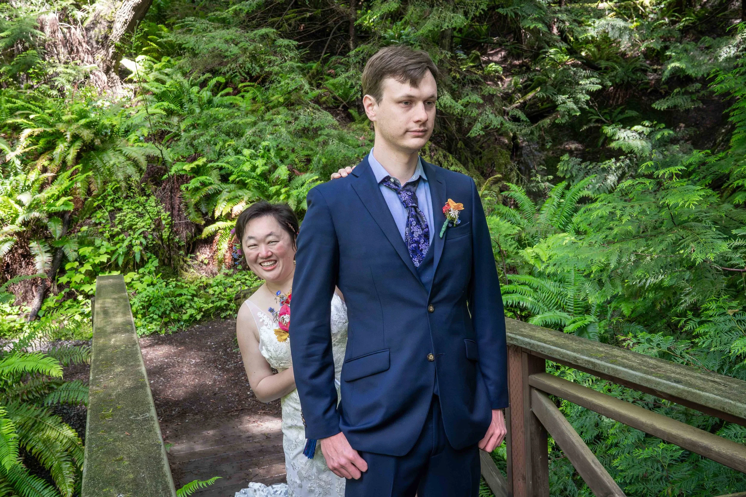 A bride in a wedding dress smiles at the camera as she comes up behind her groom and puts a hand on his shoulder during a first look ceremony in Larrabee State Park. The groom is wearing a blue suit and looking straight ahead.