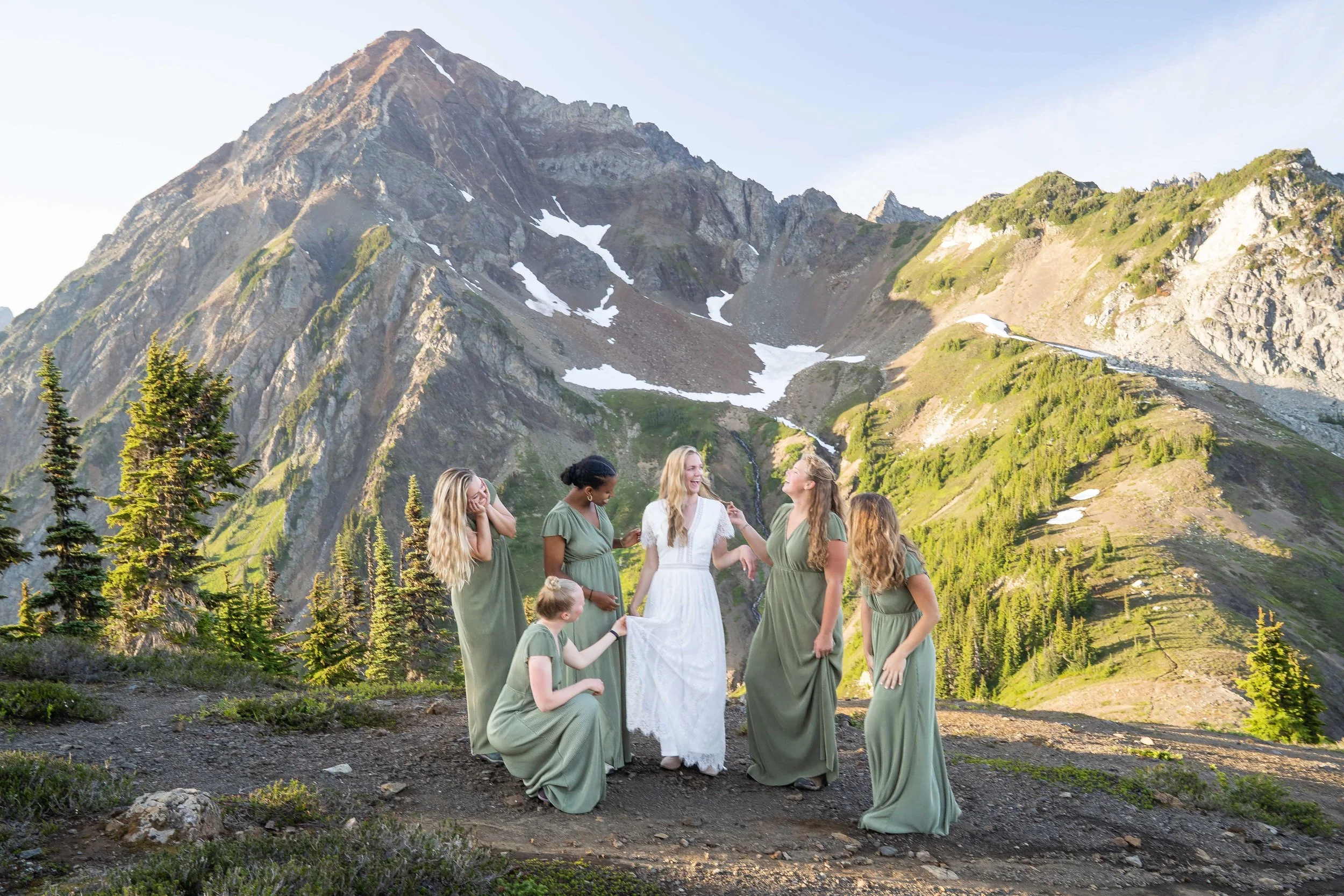 A group photo of a bride in her wedding dress being attended to by her bridesmaids in matching green dresses during a vow exchange at High Pass in the North Cascades, Washington. Mount Larrabee is directly behind the group.