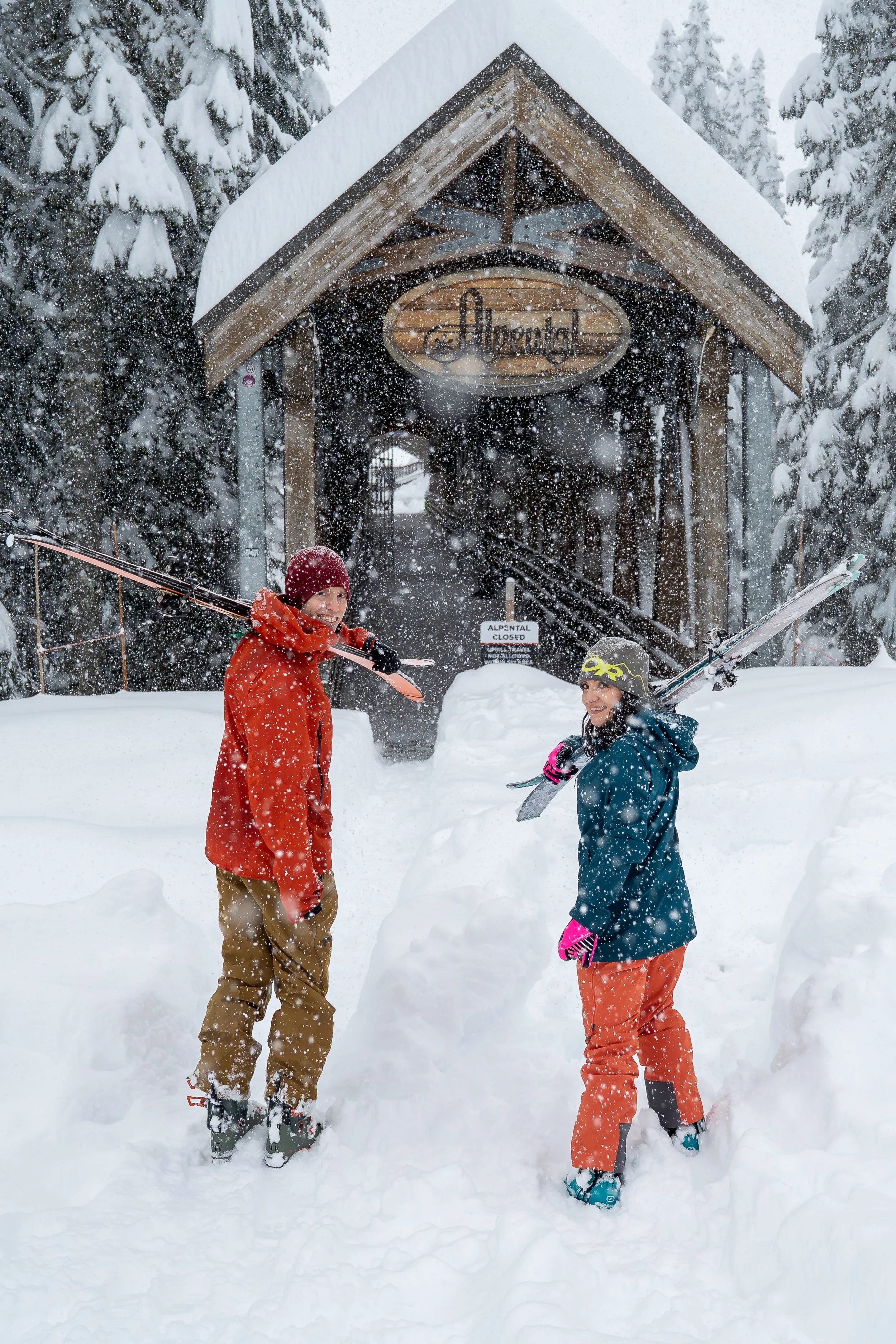 A photo of a couple in ski clothes standing in deep snow with their skis over their shoulders, looking back at the camera. The Alpental ski area sign and wooden bridge is behind them.