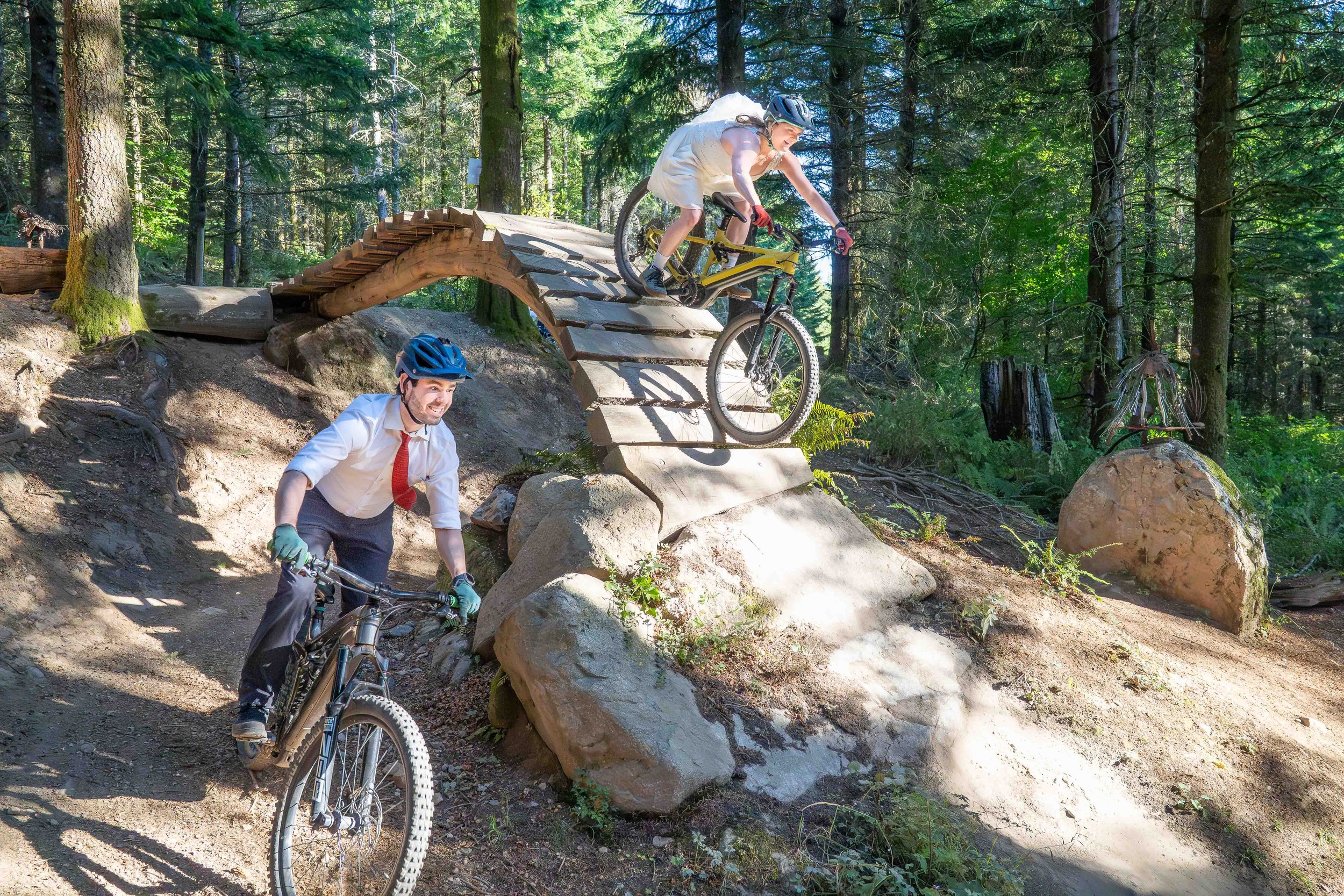 A photo of a couple in wedding clothes on mountain bikes at Galbraith Mountain in Bellingham. The bride is on a wooden bridge, while the groom is riding underneath the bridge.