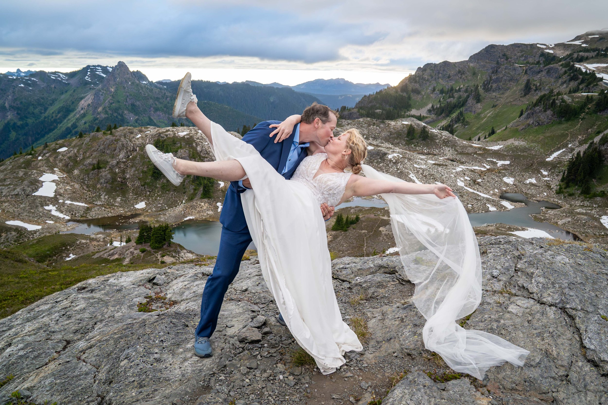 A groom holds his bride, who is wearing a wedding dress and veil, and tilts her backwards during a sunset elopement in the North Cascades. They are standing on a rocky outcrop on Yellow Aster Butte with alpine lakes and Tomyhoi Peak behind them.