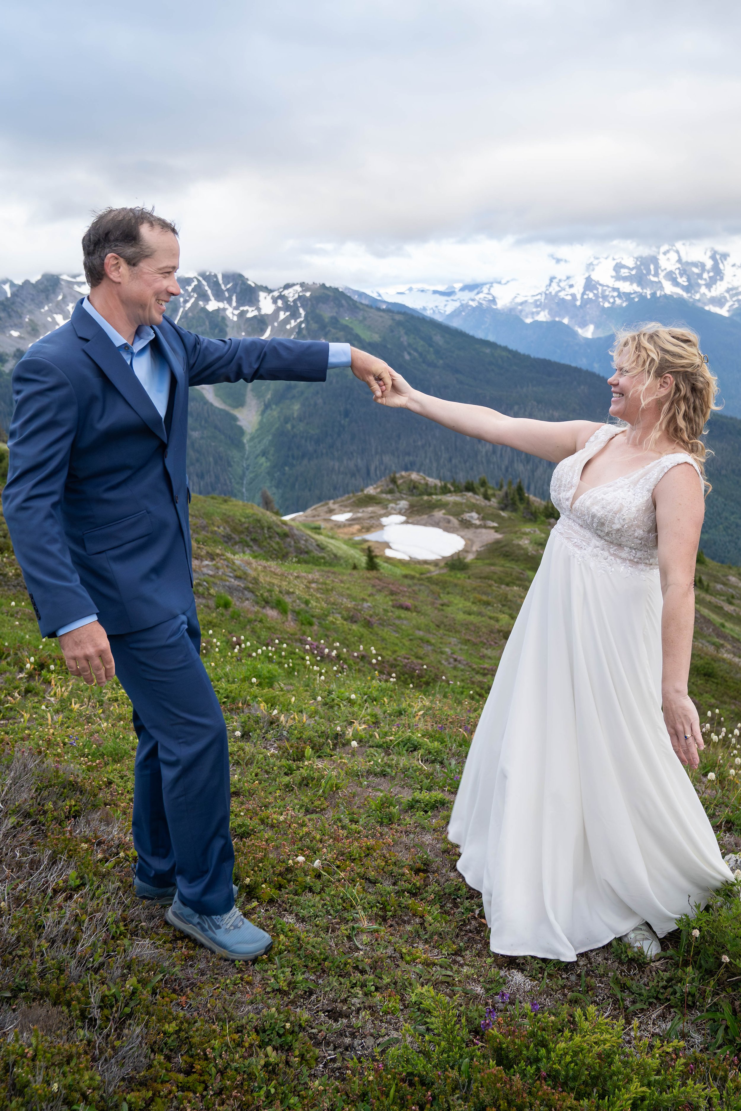 A newly married bride and groom hold their first dance in a subalpine meadow on Yellow Aster Butte at sunset during their North Cascades elopement. Mt. Shuksan is partly visible through the clouds behind them.
