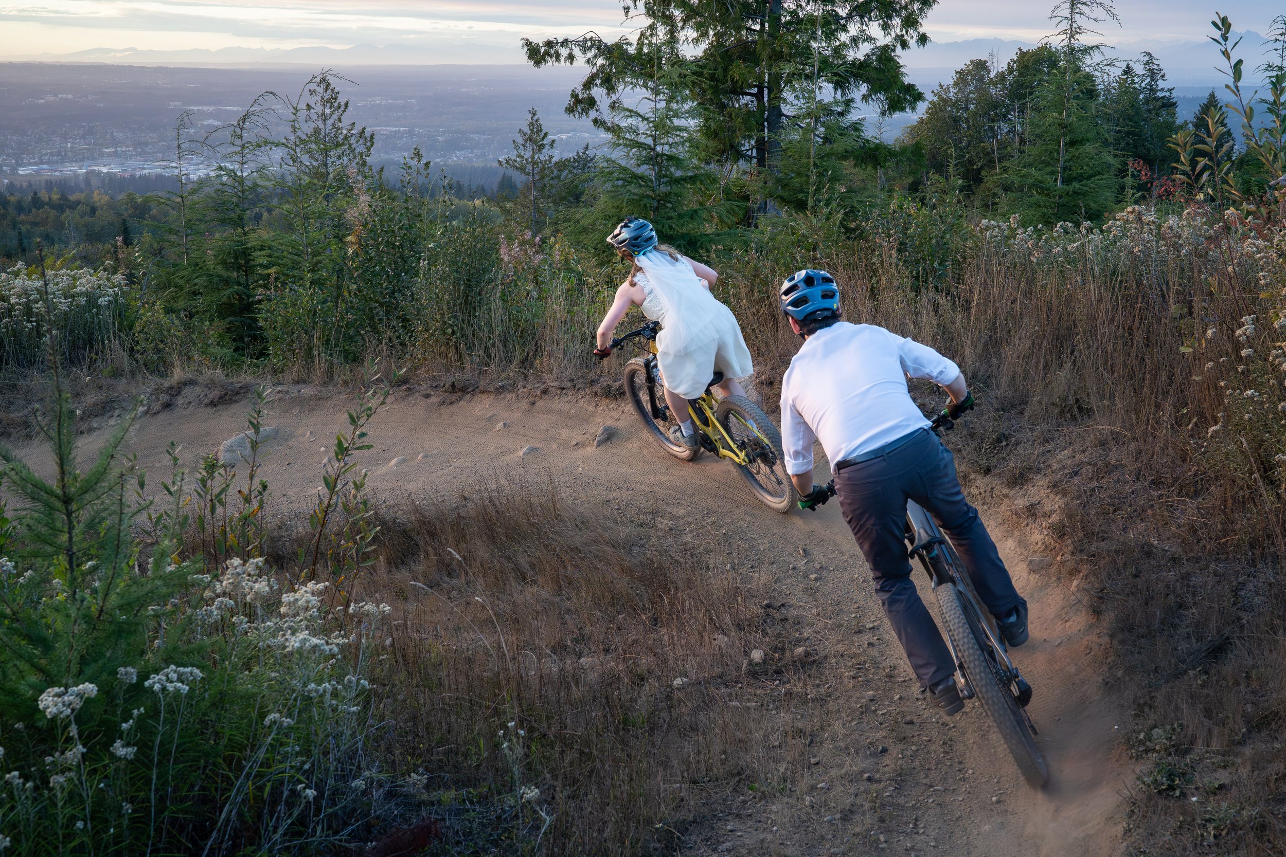 A bride and groom riding downhill at sunset on SST at the end of their Galbraith mountain bike elopement in Bellingham. The couple is in wedding clothes and the city of Bellingham is visible in the background.