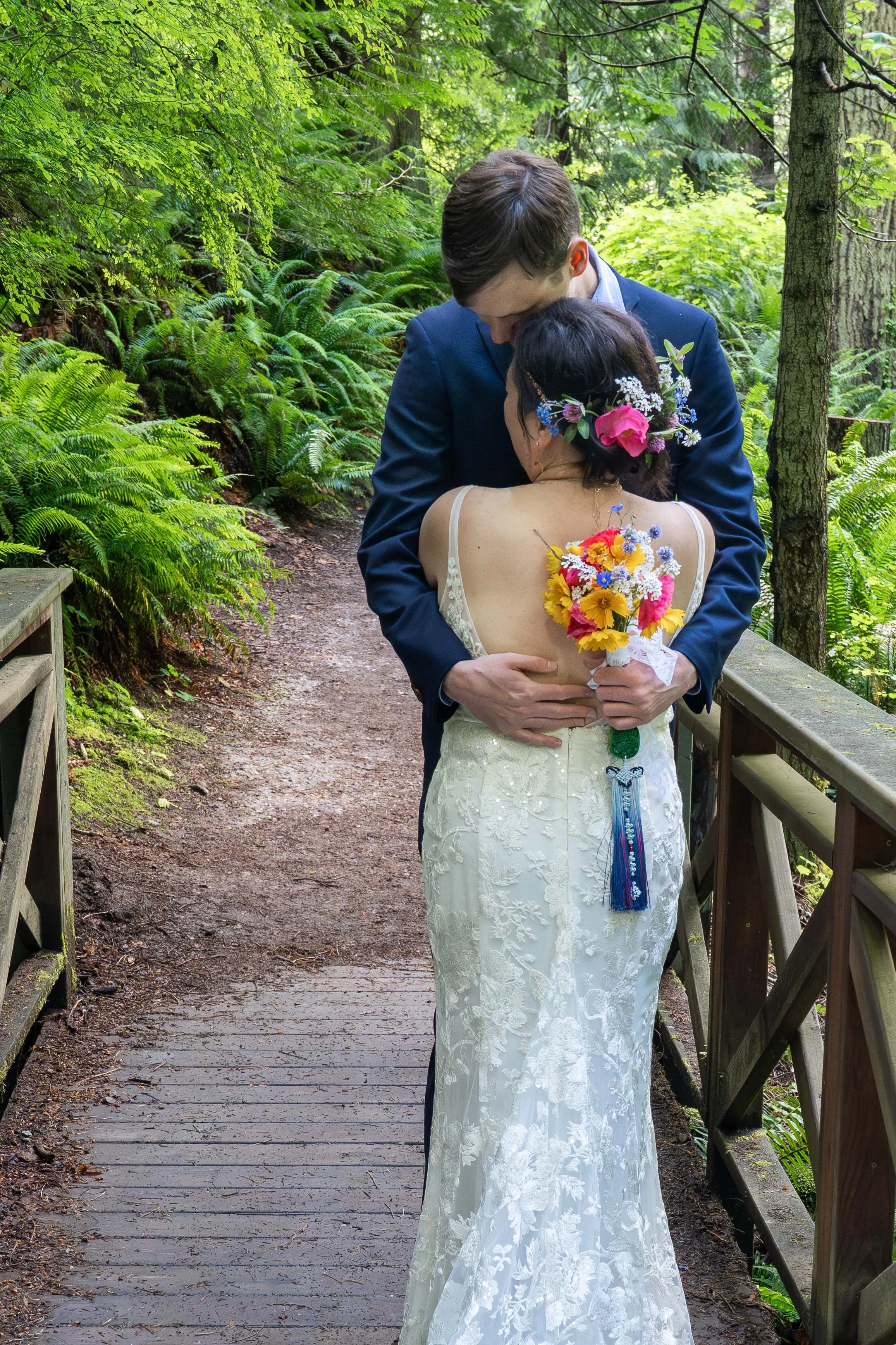 A bride and groom embrace after their first look on a wooden bride surrounded by lush ferns in Larrabee State Park, just before their elopement ceremony. The bride's back is to the camera and the groom is holding her bouquet.