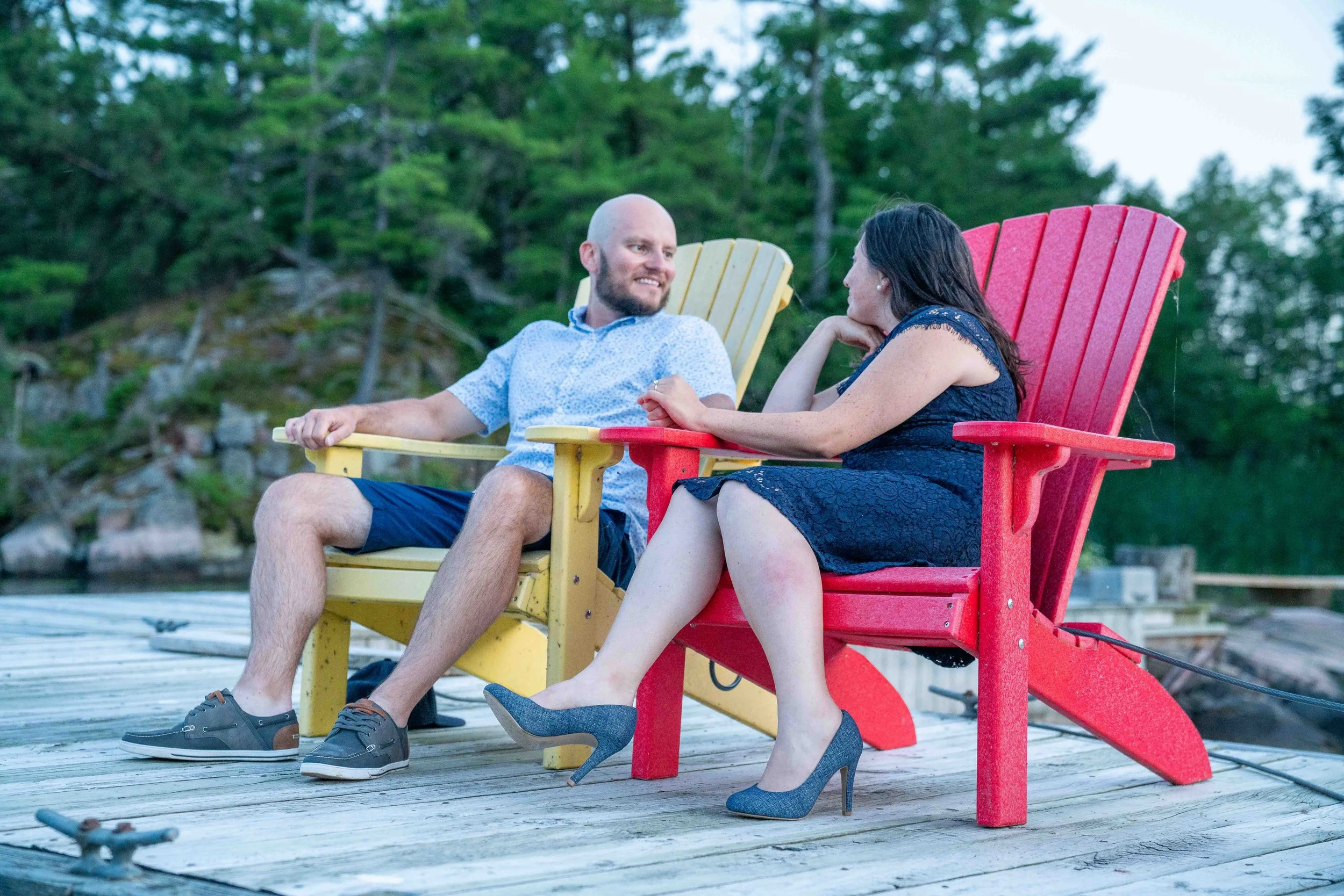 A couple in formal wear sitting on yellow and red Adirondack chairs on a dock with trees in the background.