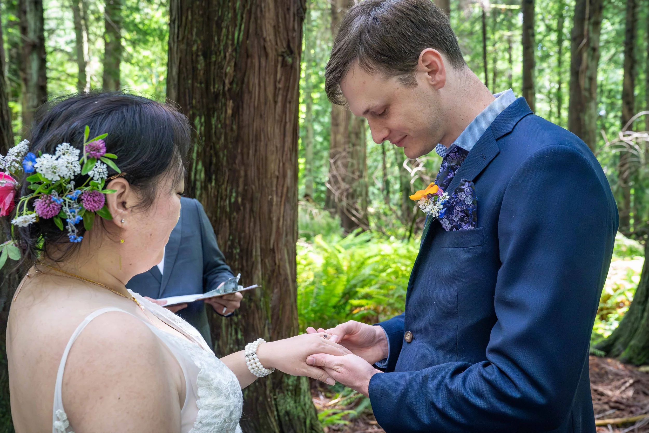 A groom in a blue suit putting a ring on his bride's finger during their adventure wedding ceremony in Larrabee State Park.