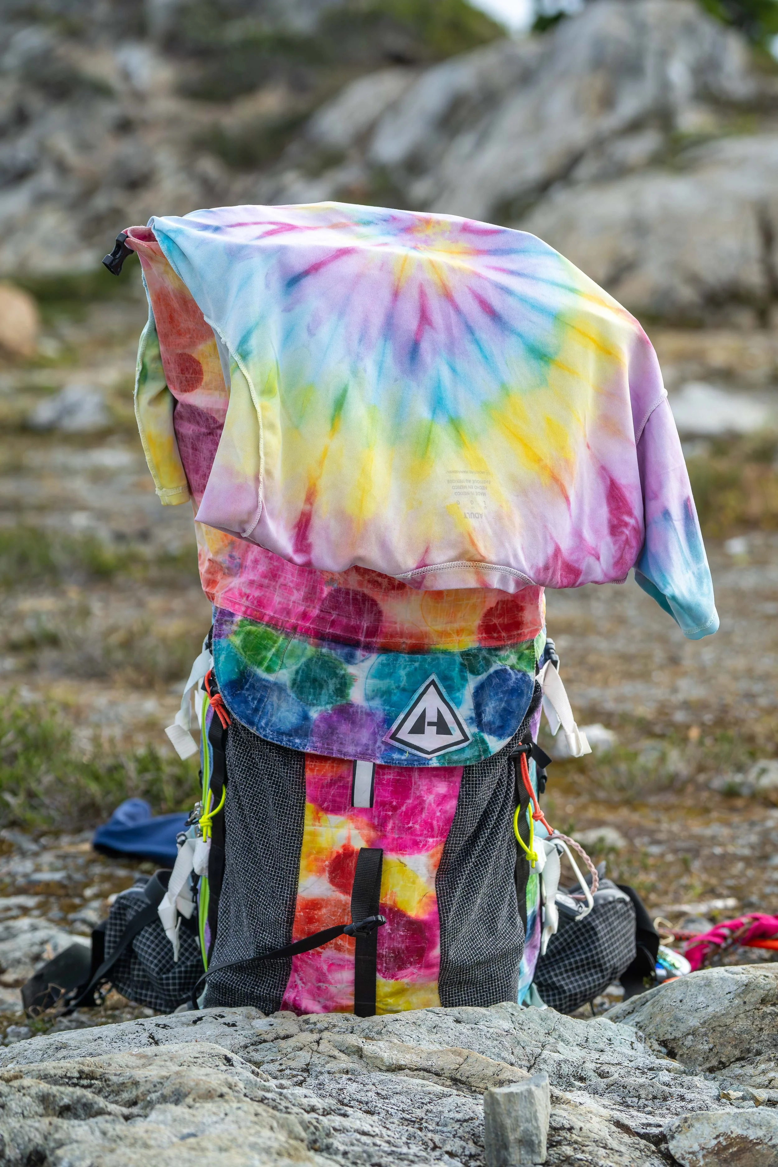 A brightly colored, tie-dye backpack with a tie-dye t-shirt hanging on top of it to dry. The pack is sitting in the middle of a rocky alpine meadow in the North Cascades.