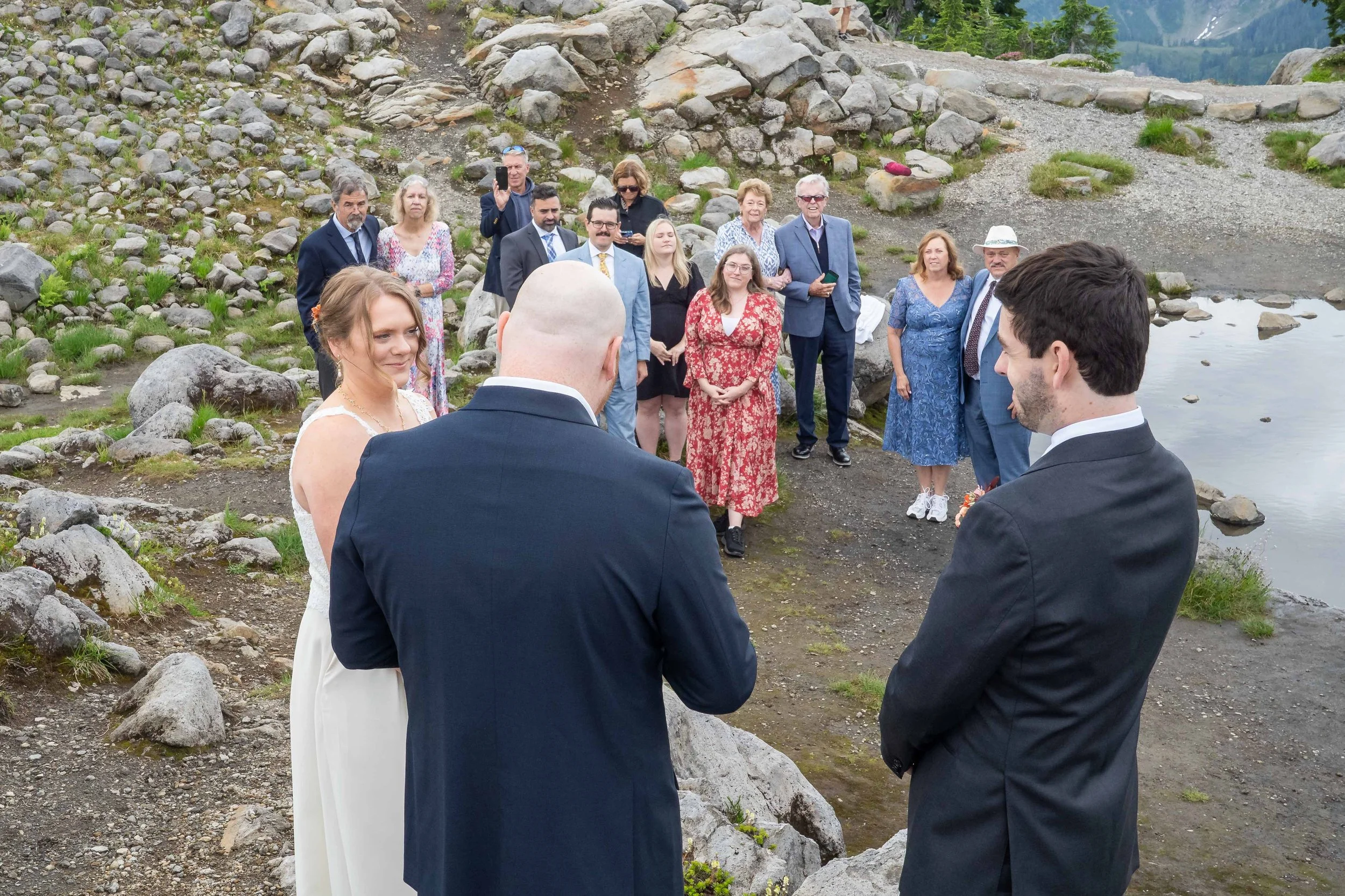 Photo of a wedding ceremony in an alpine meadow at Artist Point with family looking on. The photo is taken from behind the officiant, with the bride and groom visible and their family looking on.