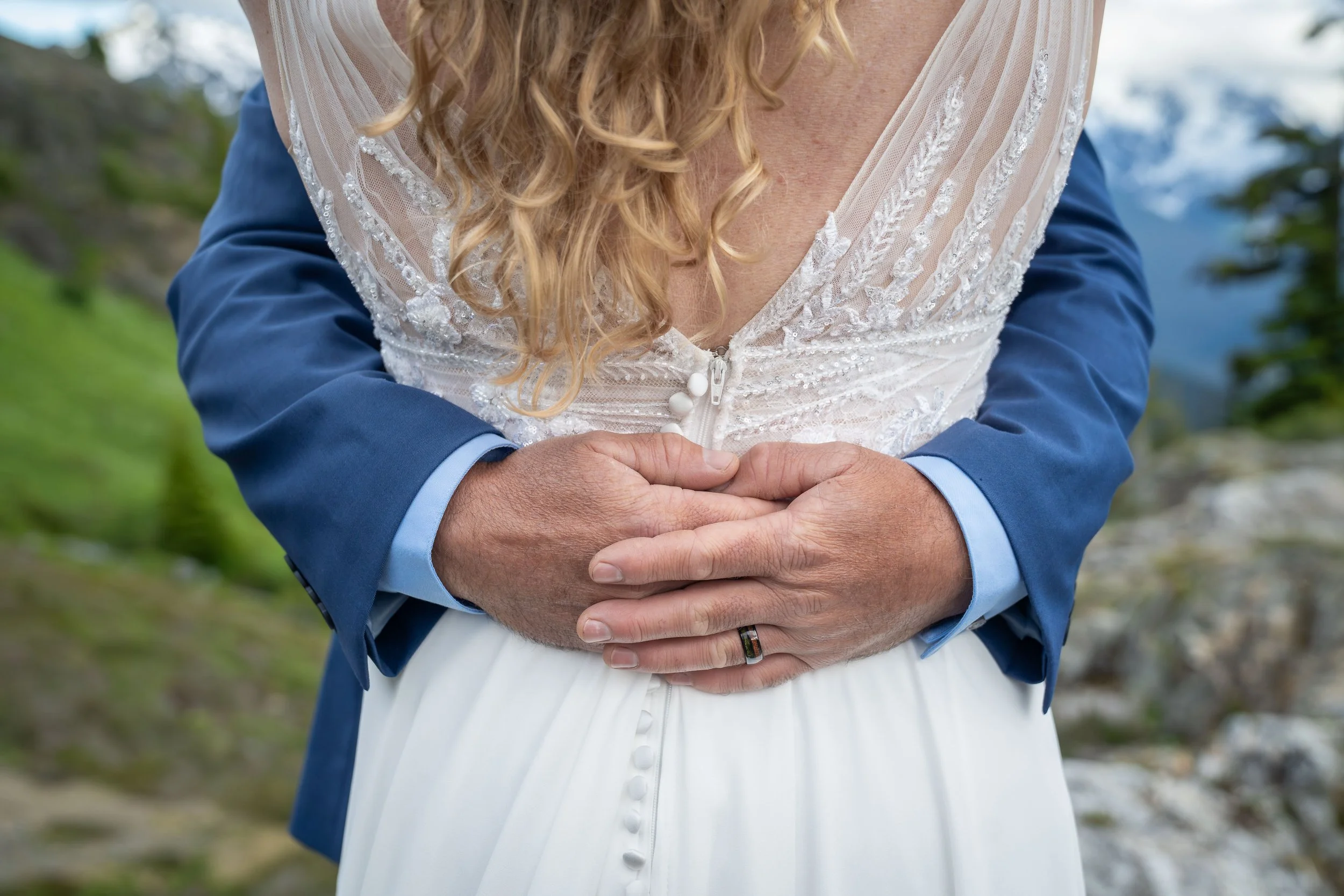 A groom's hands are wrapped around his bride's waist so that his wedding ring is clearly visible during their North Cascades elopement ceremony. The photo is taken from behind the bride, showing her dress, and Mt. Shuksan is visible behind them.