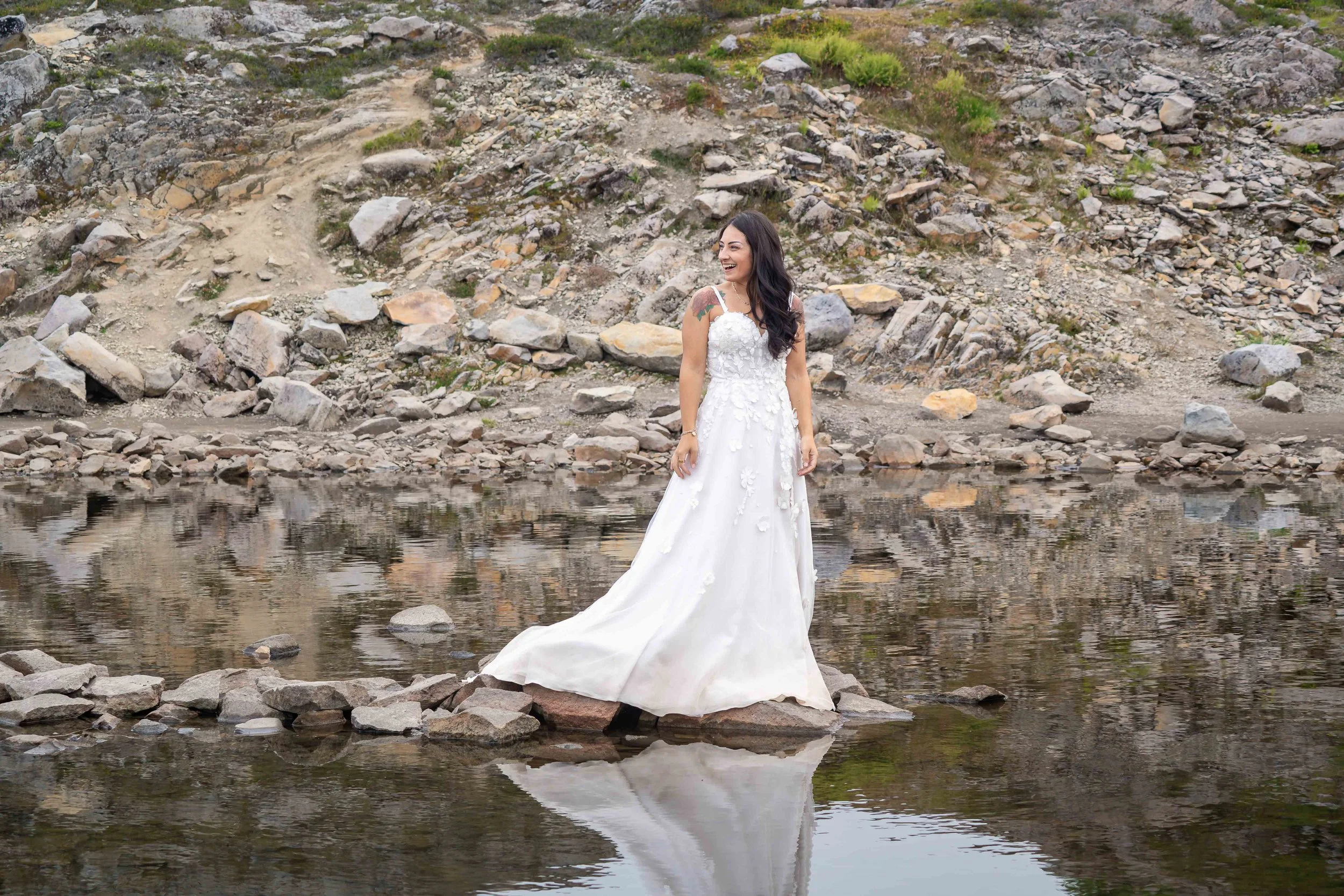 A bride in a flowing wedding dress standing on rocks in the middle of an alpine lake at Artist Point during her Mt. Baker adventure wedding. She is looking back at her groom on the shore of the lake.