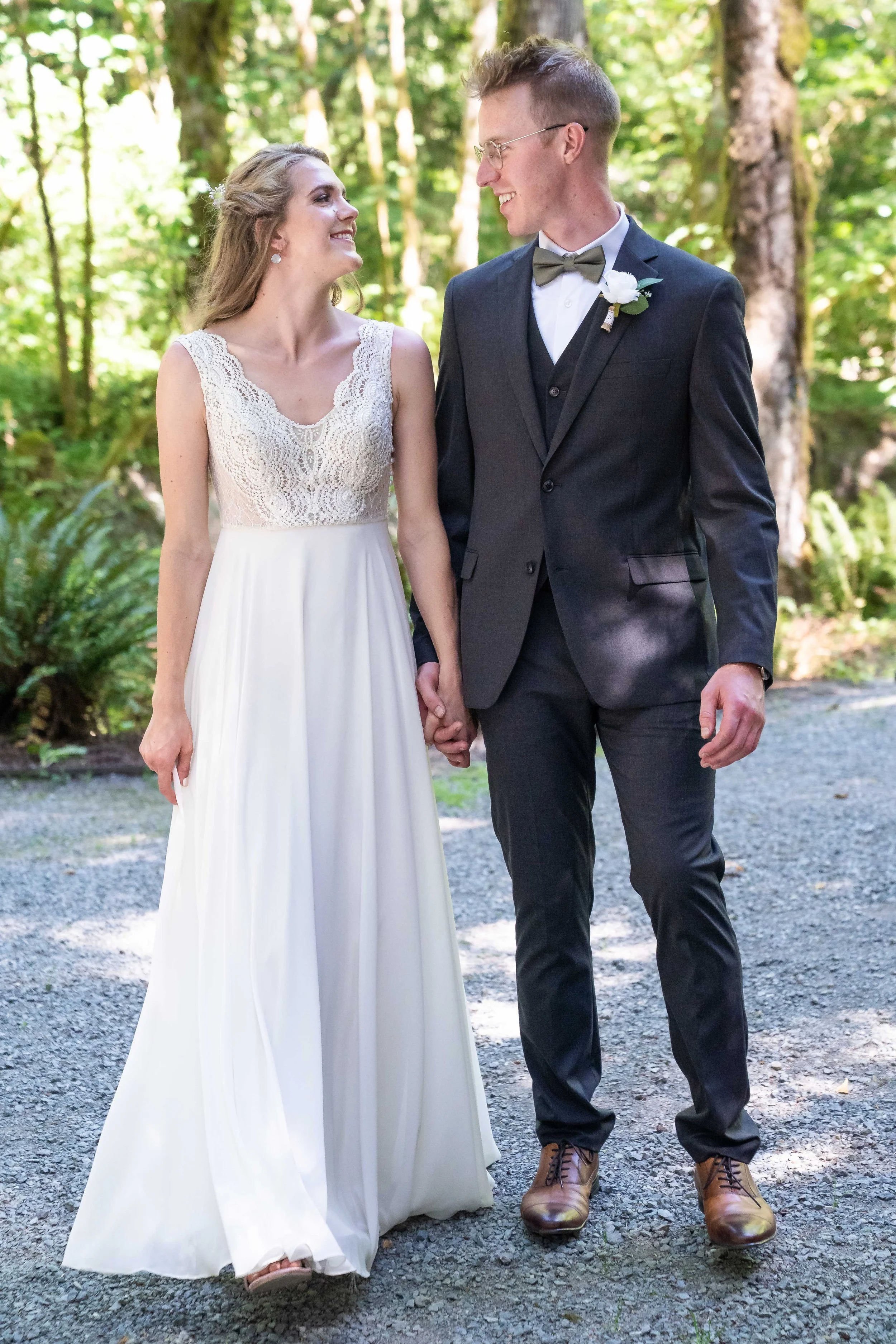 A bride and groom standing side-by-side while holding hands and looking at each other during a sunny forest wedding in Skagit County, Washington.