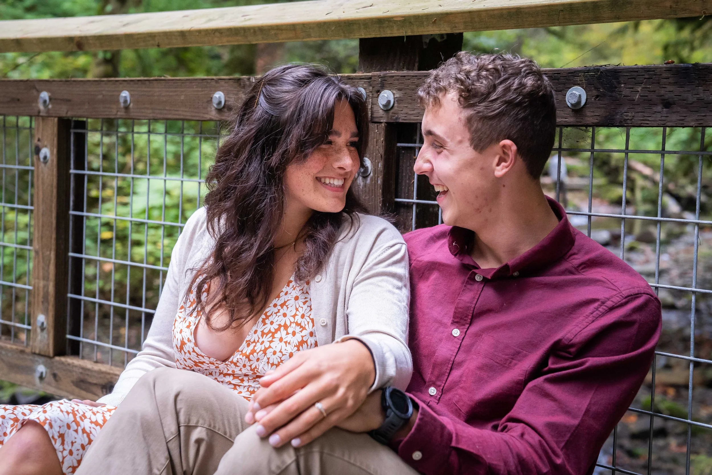A photo of a couple in a button-down shirt and dress sitting together on a wooden bridge at Arroyo Park in the Chuckanuts. The woman's engagement ring is clearly visible and there's a lush green forest behind them.