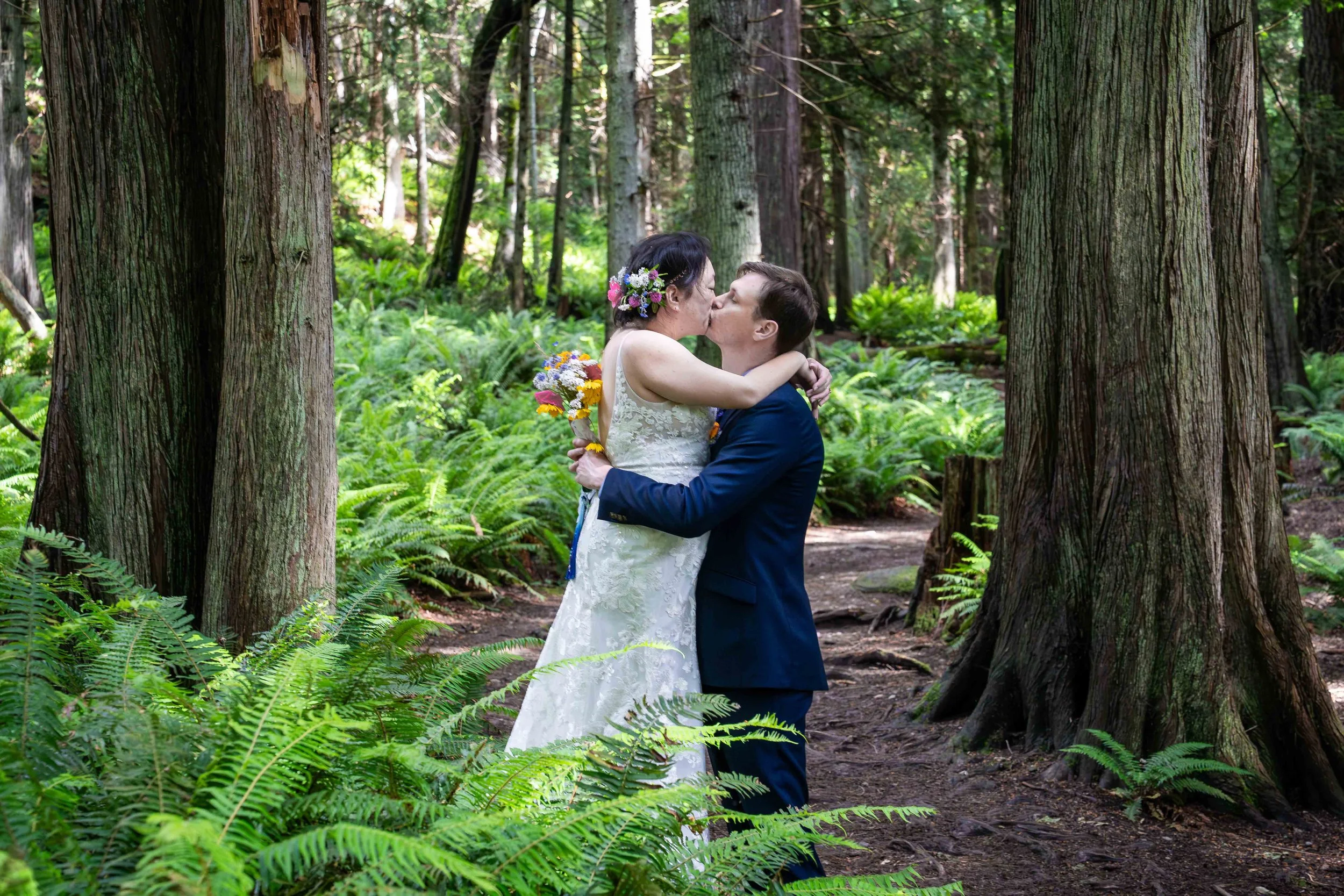 A bride and groom kiss after being married during a Larrabee State Park elopement ceremony. They are on the Fragrance Lake Trail, surrounded by old growth trees and bright green ferns.
