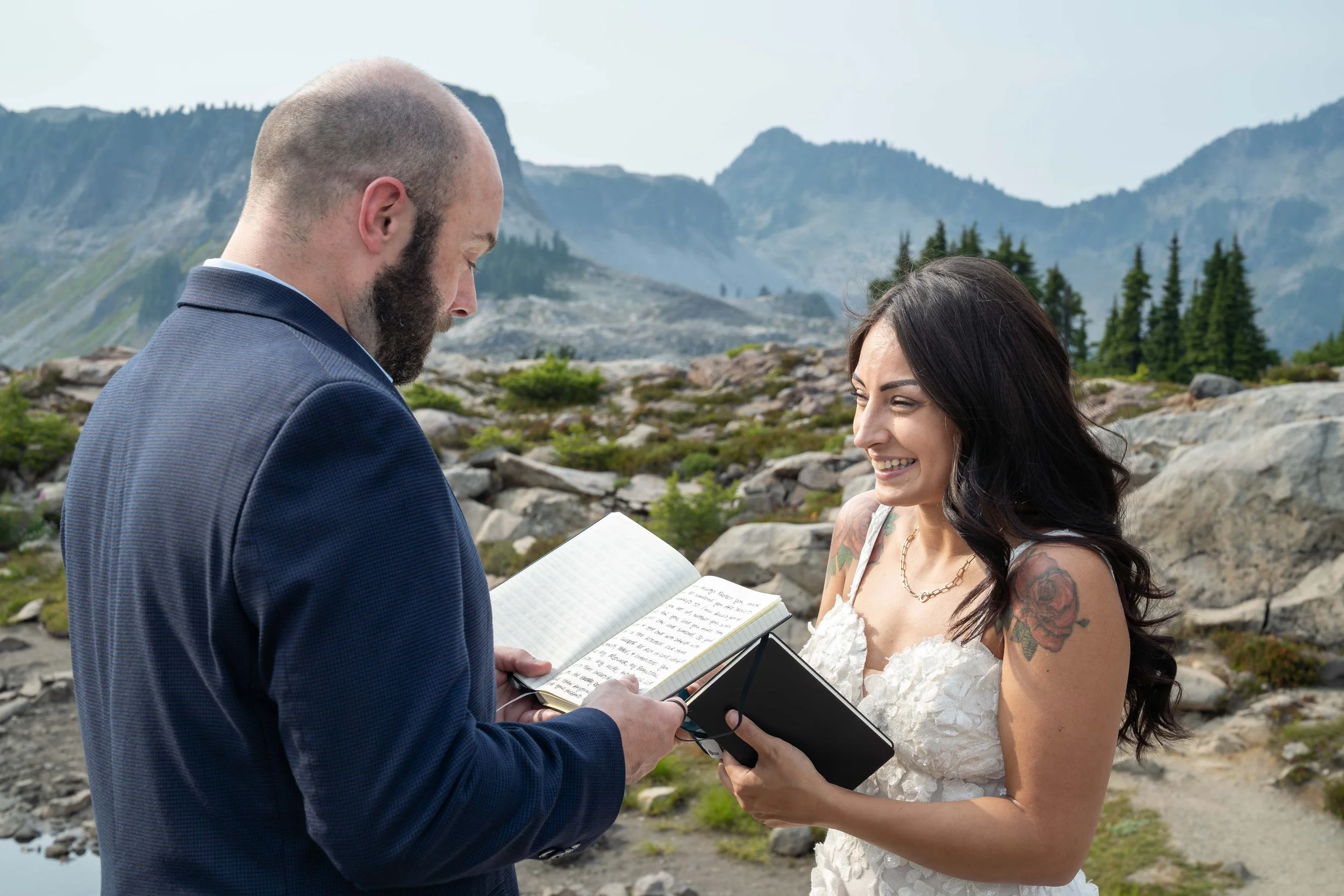A groom reading his handwritten vows from a vow book during an elopement ceremony at Artist Point. His bride is smiling and crying, and a subalpine meadow and Table Mountain are visible behind them.