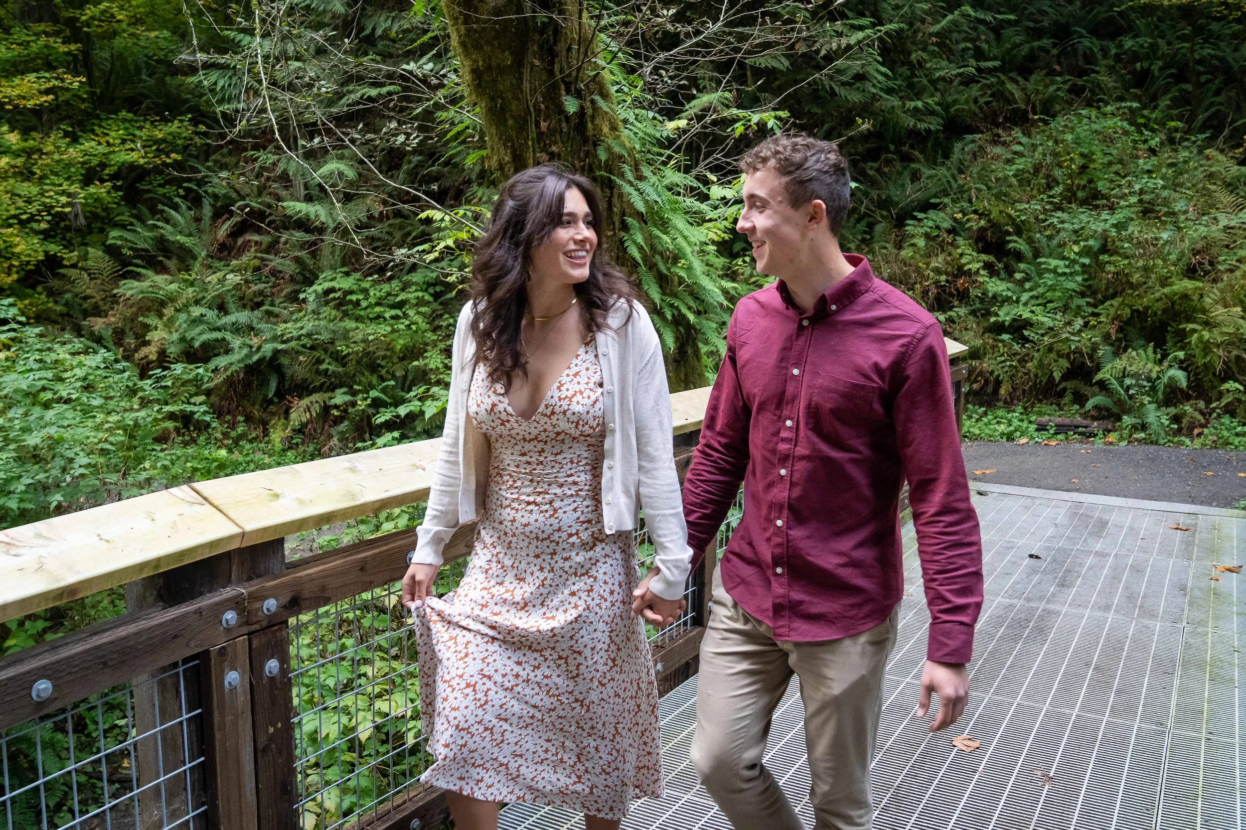 A photo of a couple walking together, holding hands and smiling at each other, on a wooden bridge at Arroyo Park in Bellingham. The woman is wearing a sundress and the man is wearing a red button-down shirt.