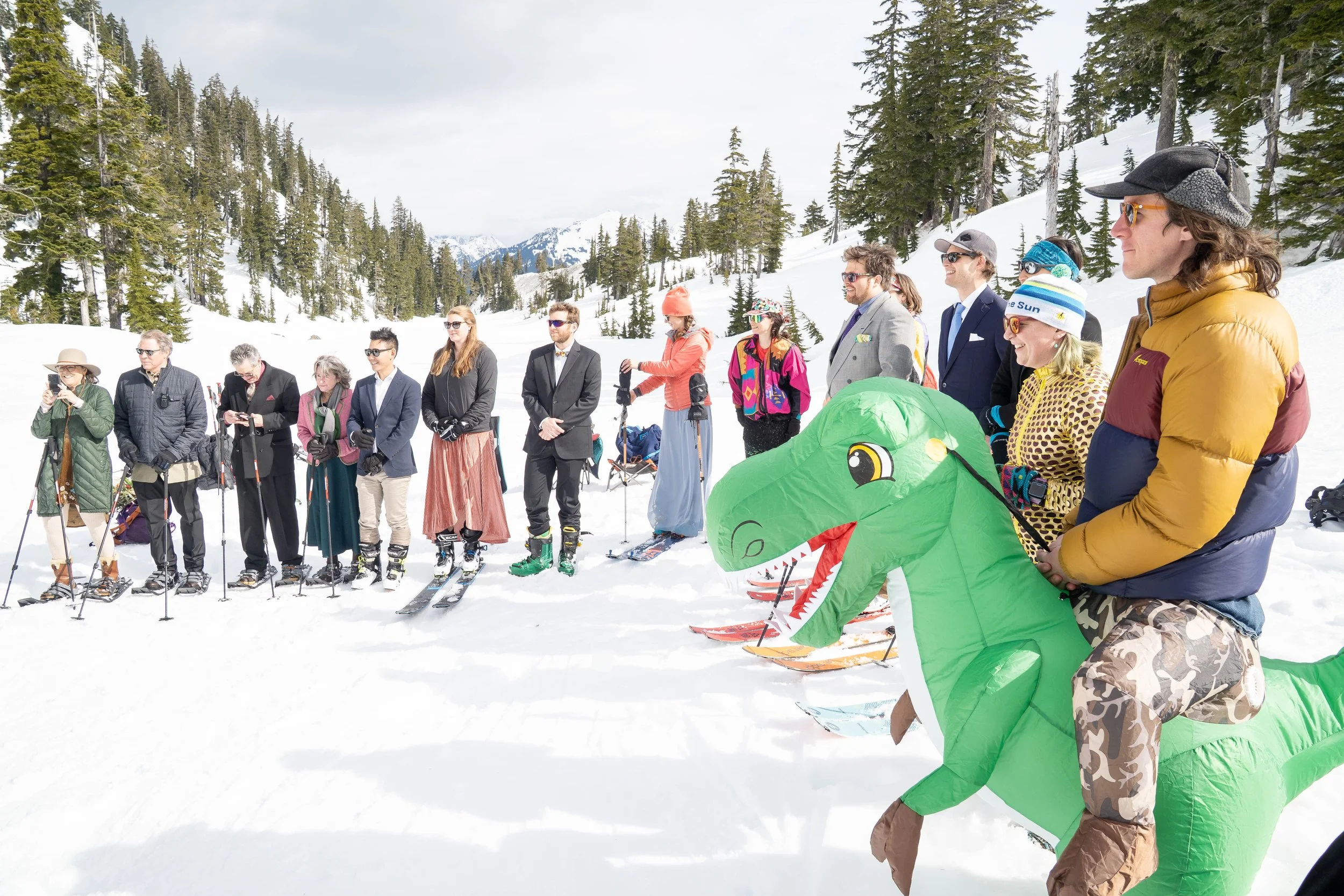 A photo of assembled friends and family in the snow at Heather Meadows watching a wedding ceremony. One person in the foreground is wearing a dinosaur costume and a puffy jacket.