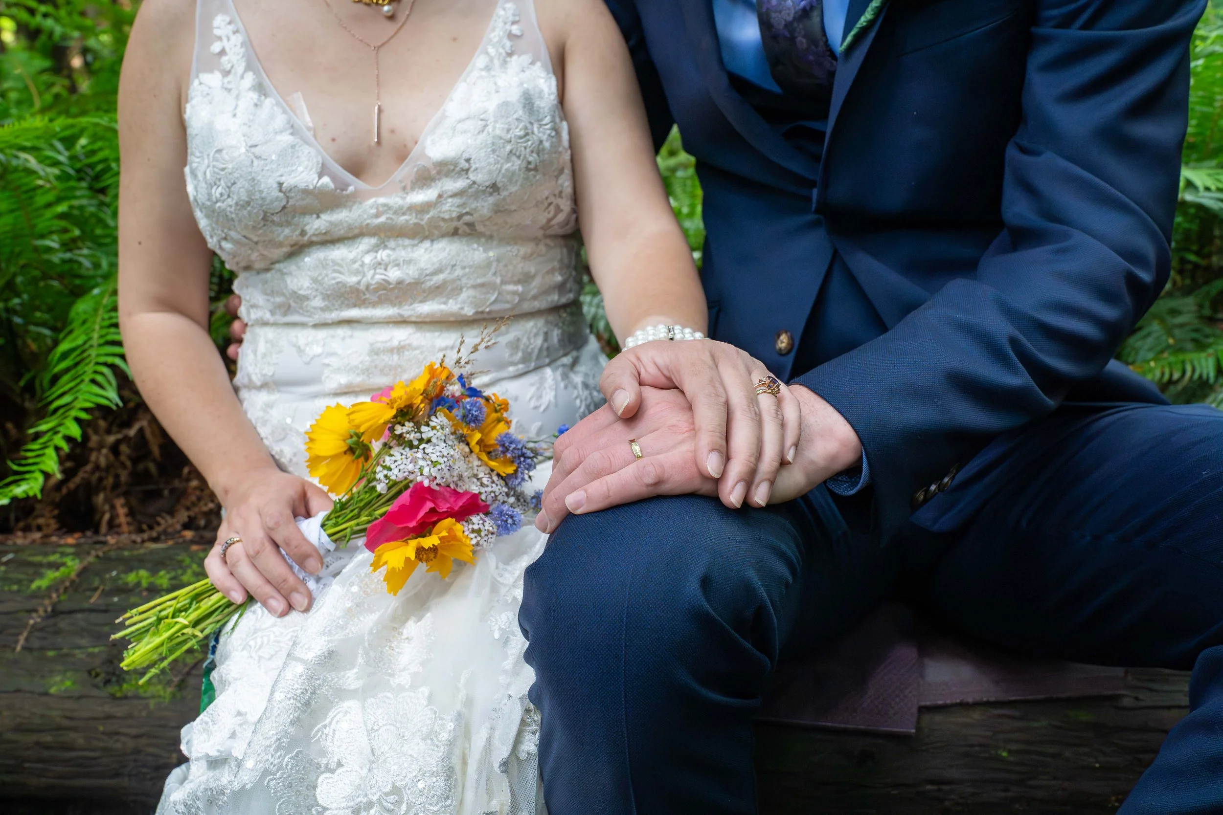 A bride and groom sit on a log in Larrabee State Park and hold hands with their wedding rings clearly visible.