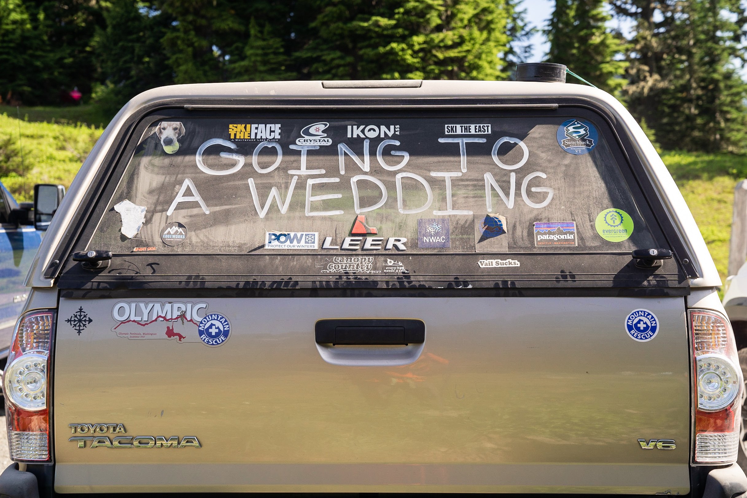 A Toyota Tacoma with a bed topper covered in outdoor-related stickers and white paint that says "Going to a Wedding" parked at a trailhead in the North Cascades.