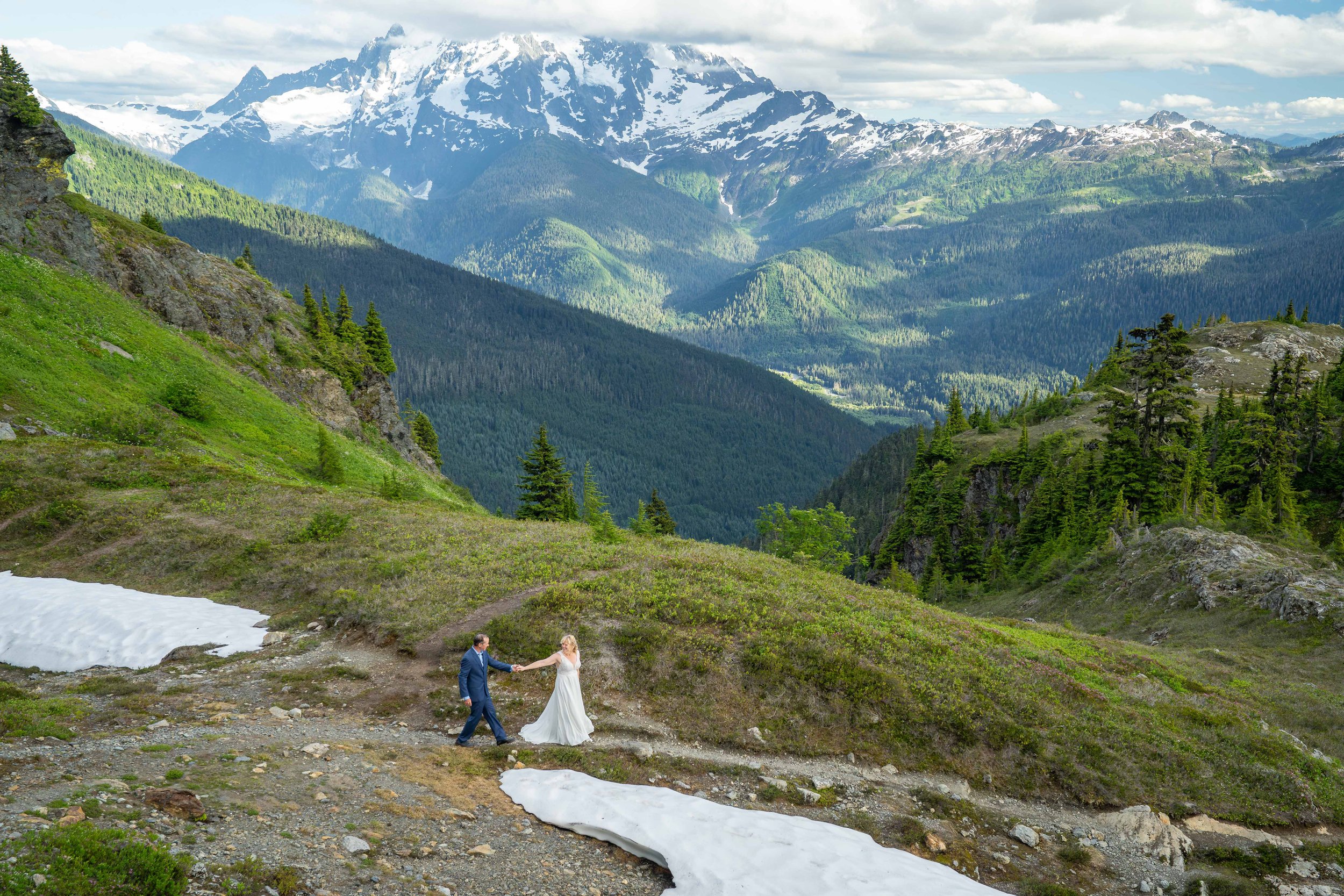 A bride leading her groom by the hand down a rocky trail through an alpine meadow of wildflowers in the North Cascades, with Mt. Shuksan and the Nooksack Valley visible behind them.