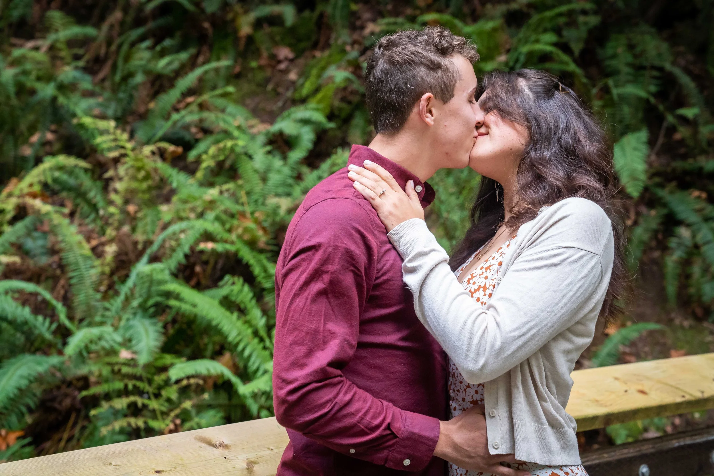 A photo of a couple in a button-down shirt and dress standing together and kissing on a wooden bridge at Arroyo Park in the Chuckanuts. The woman's engagement ring is clearly visible and there's a lush green forest behind them.