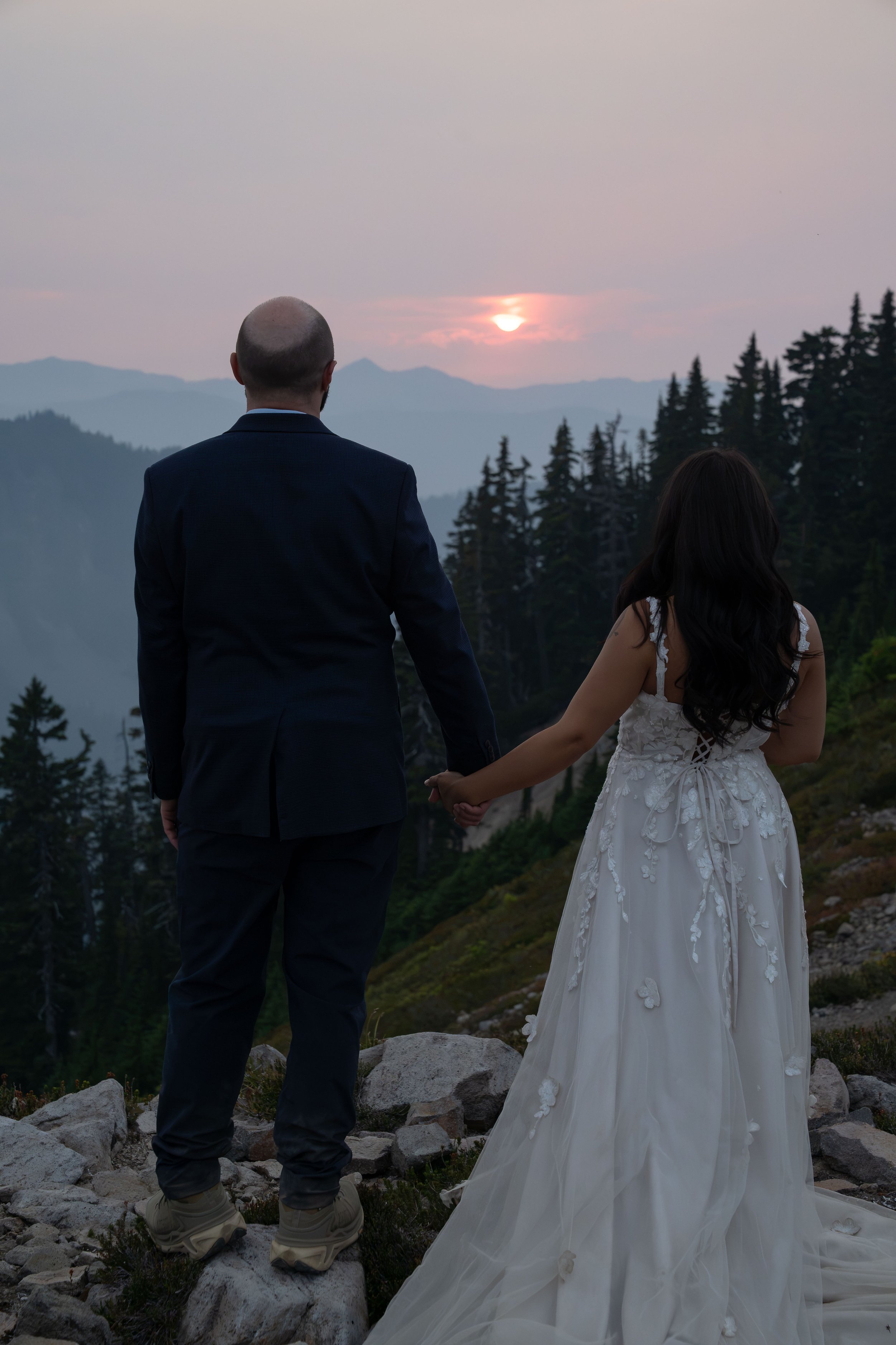 A silhouette photo of a bride and groom standing together watching the sunset from Ptarmigan Ridge during their Mt. Baker elopement. They are standing with their backs to the camera and the sun is a vibrant pink thanks to haze from wildfire smoke.